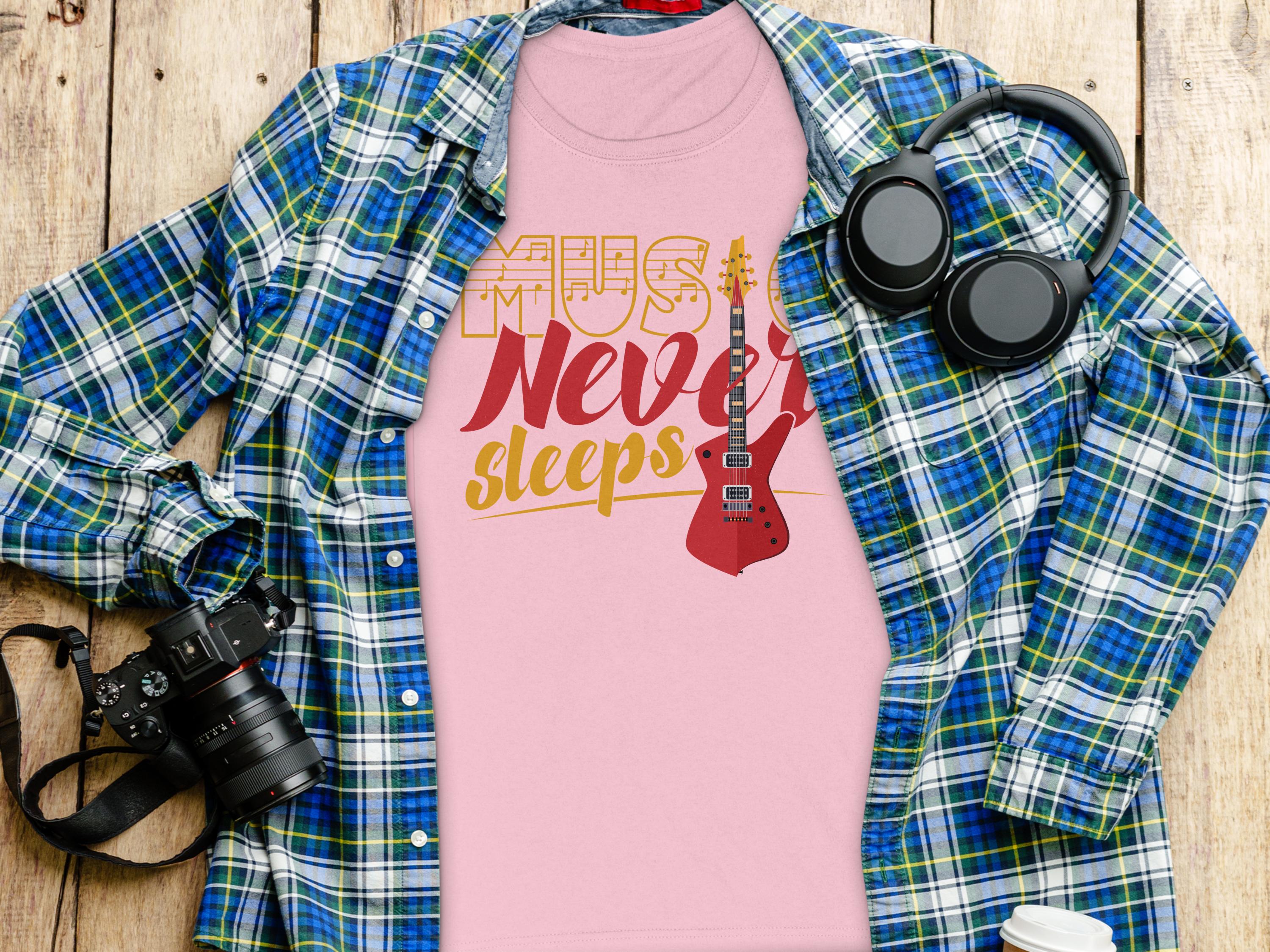 A unique pink Garment Graphics T-shirt featuring Music Never Sleeps and a guitar graphic is displayed on a wooden surface, complemented by a blue and green plaid shirt, headphones, a camera, and a white takeaway coffee cup—perfect for guitar T-shirt enthusiasts.