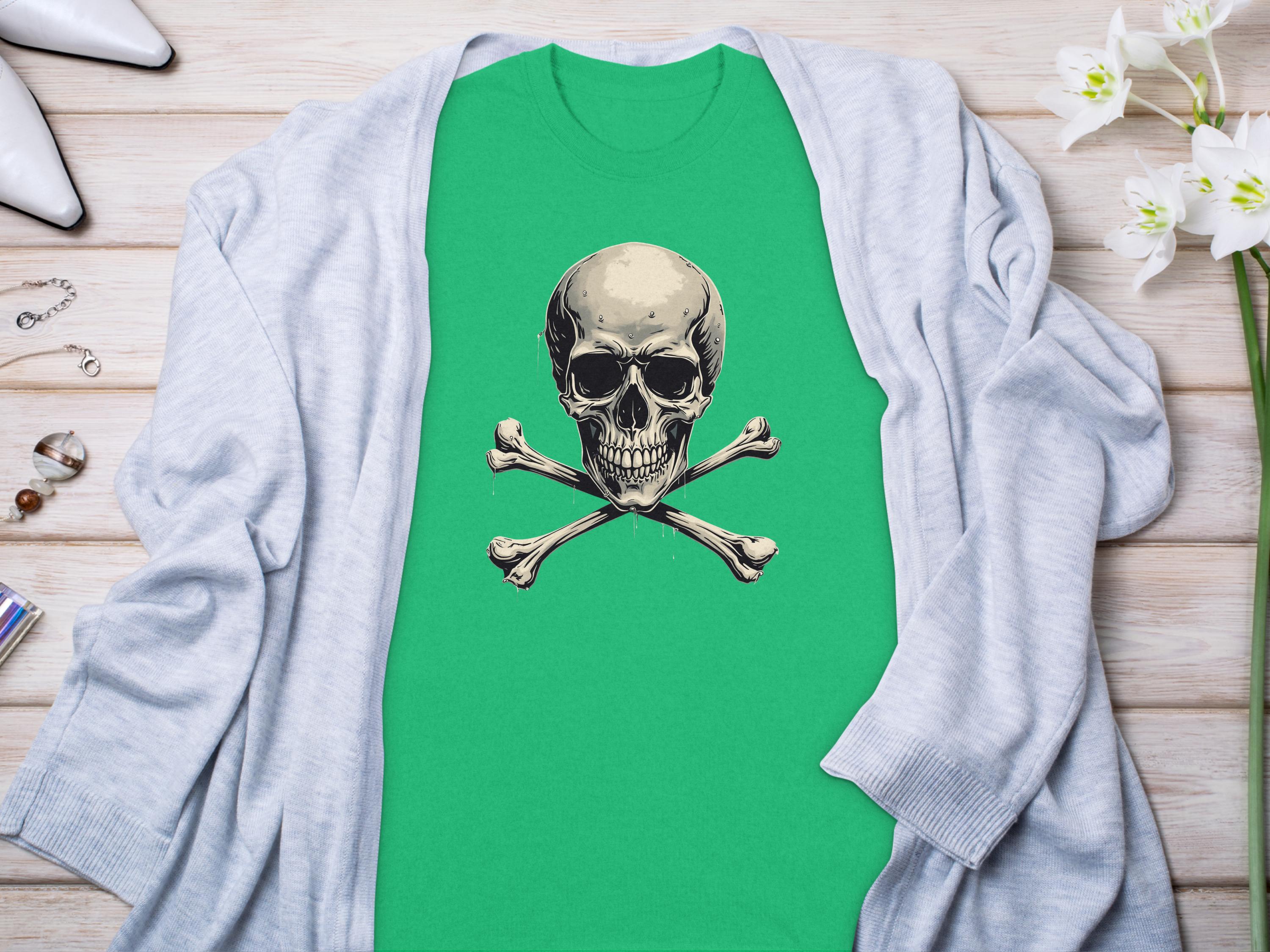 A green Garment Graphics graphic t-shirt with a striking skull and crossbones design is laid on wood. Its stylishly paired with a gray cardigan, white high heels, jewelry, and delicate white flowers for an edgy fashion look.