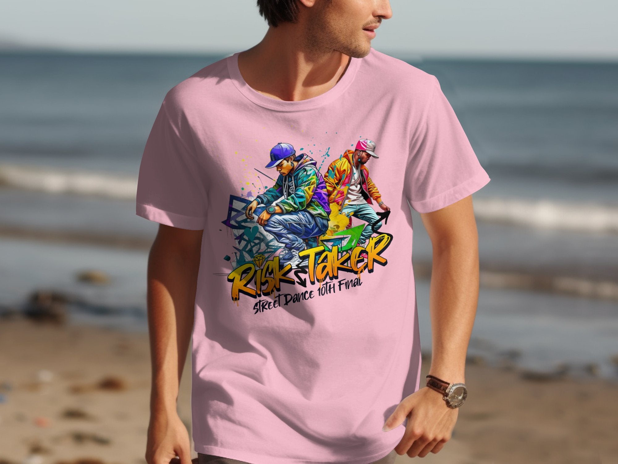A person sports a Garment Graphics pink t-shirt showcasing an artistic design of two dancers, one blue and the other pink, alongside the phrases Risk Taker and Street Dance with Flava, creating a dynamic nod to street dance culture against the ocean backdrop.