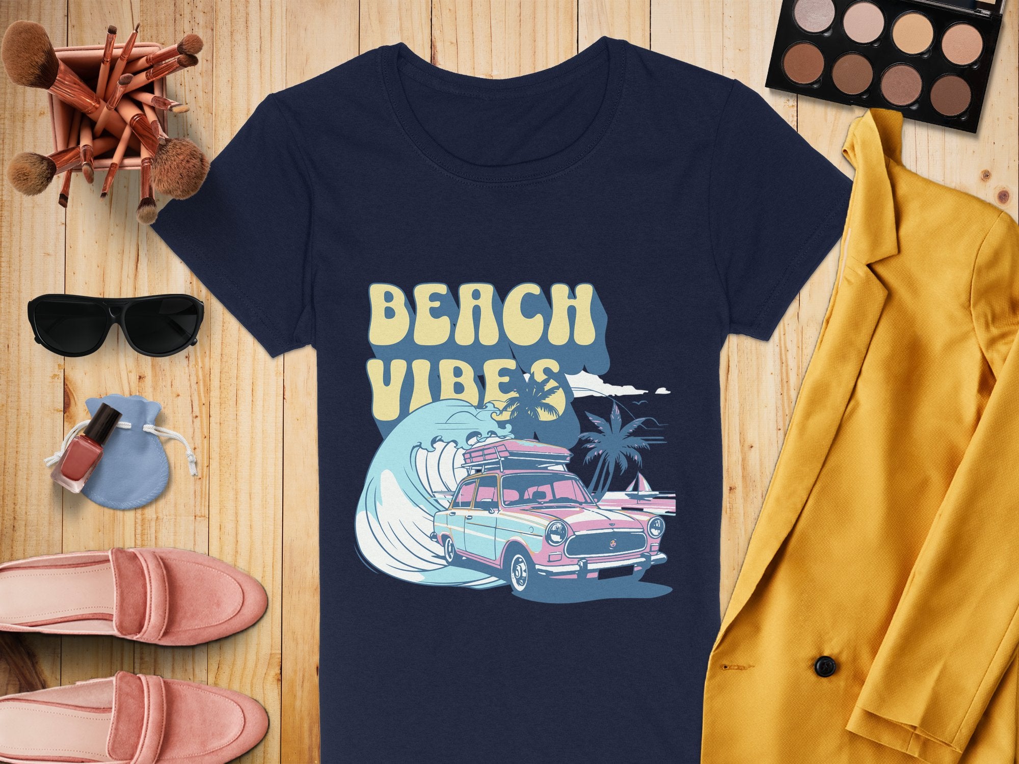 The flat lay features a Garment Graphics navy t-shirt with a surf design—Beach Vibes with a car, surfboard, and palm trees—accompanied by a yellow jacket, pink loafers, sunglasses, makeup brushes, nail polish, and an eyeshadow palette—ideal for pairing with retro t-shirts.