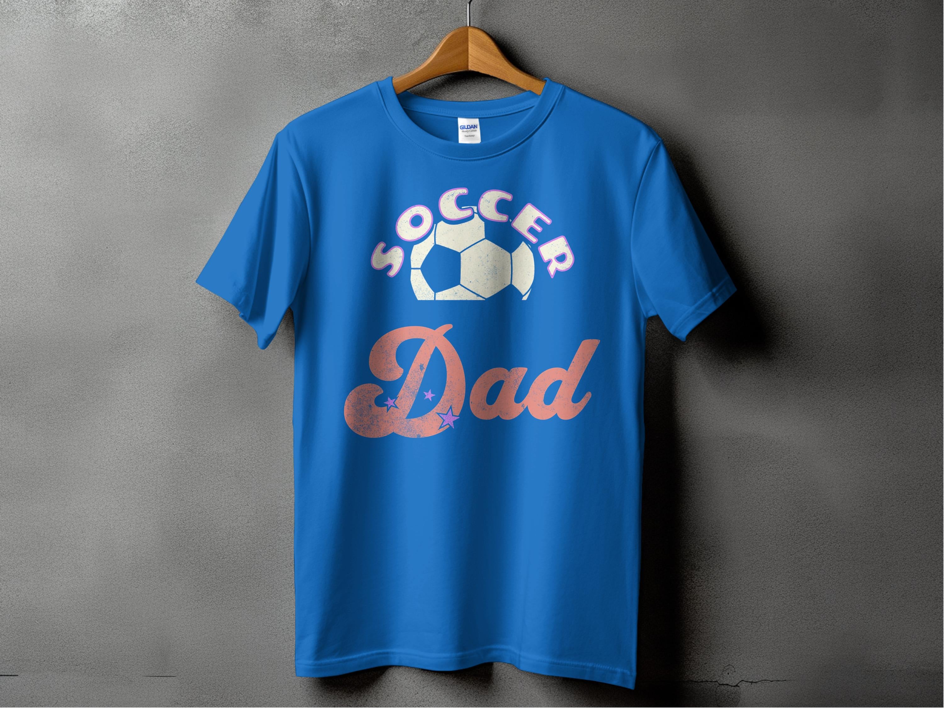 A blue graphic T-shirt from Garment Graphics hangs on a wooden hanger against a dark wall, showcasing the text SOCCER Dad with a soccer ball graphic above Dad.