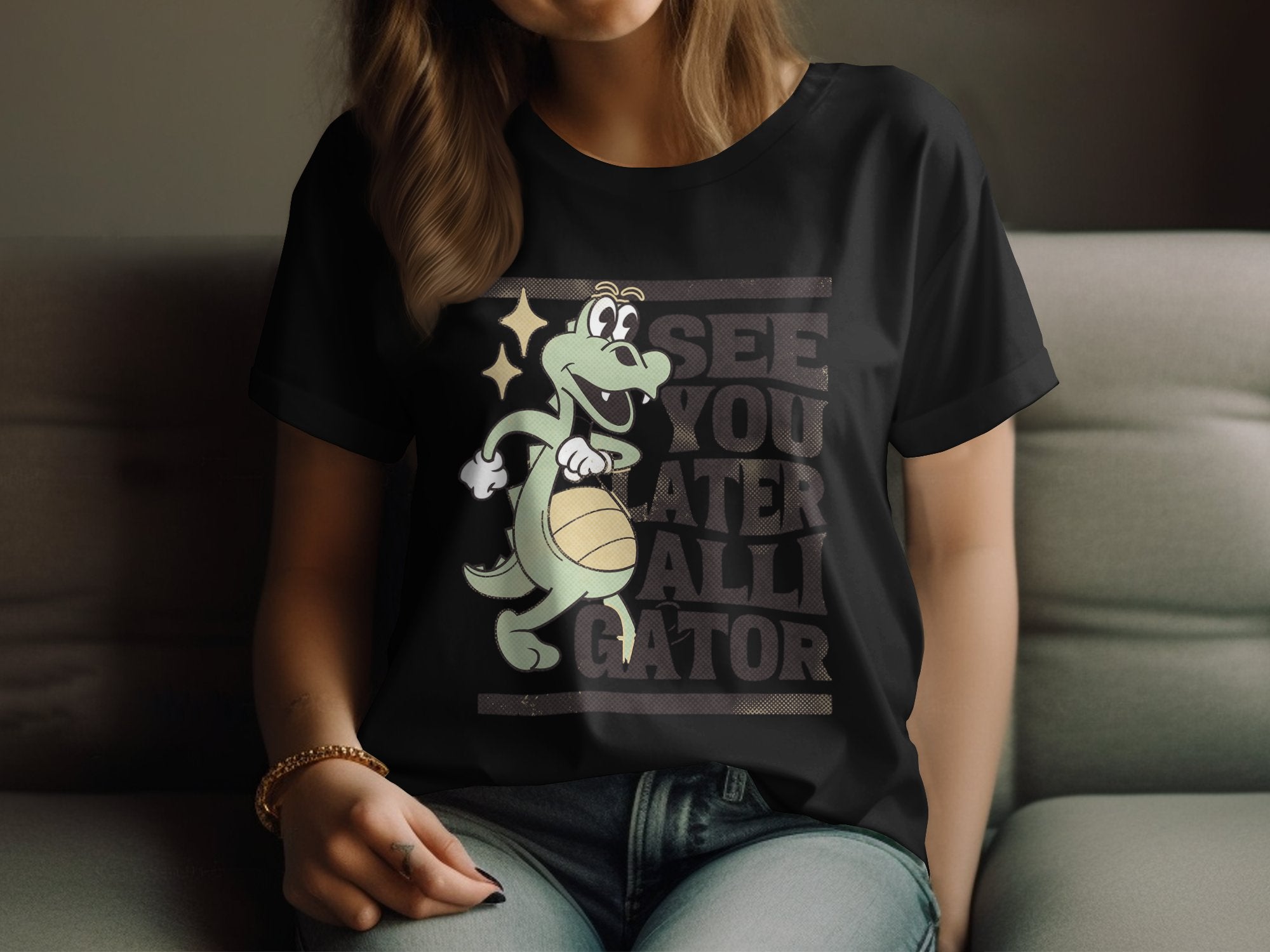 A person relaxes on a sofa wearing a Garment Graphics black womens T-shirt featuring a cartoon alligator holding a basketball with the phrase See you later alligator.