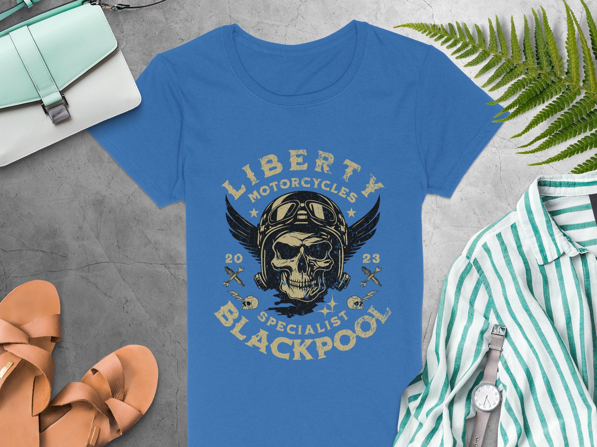 Garment Graphics blue T-shirt features a skull with wings, goggles, and a helmet design that reads Liberty Motorcycles Specialist Blackpool 2023. Ideal for pairing with sandals, a striped shirt, watch, purse, and fern leaf for a relaxed Blackpool vibe.