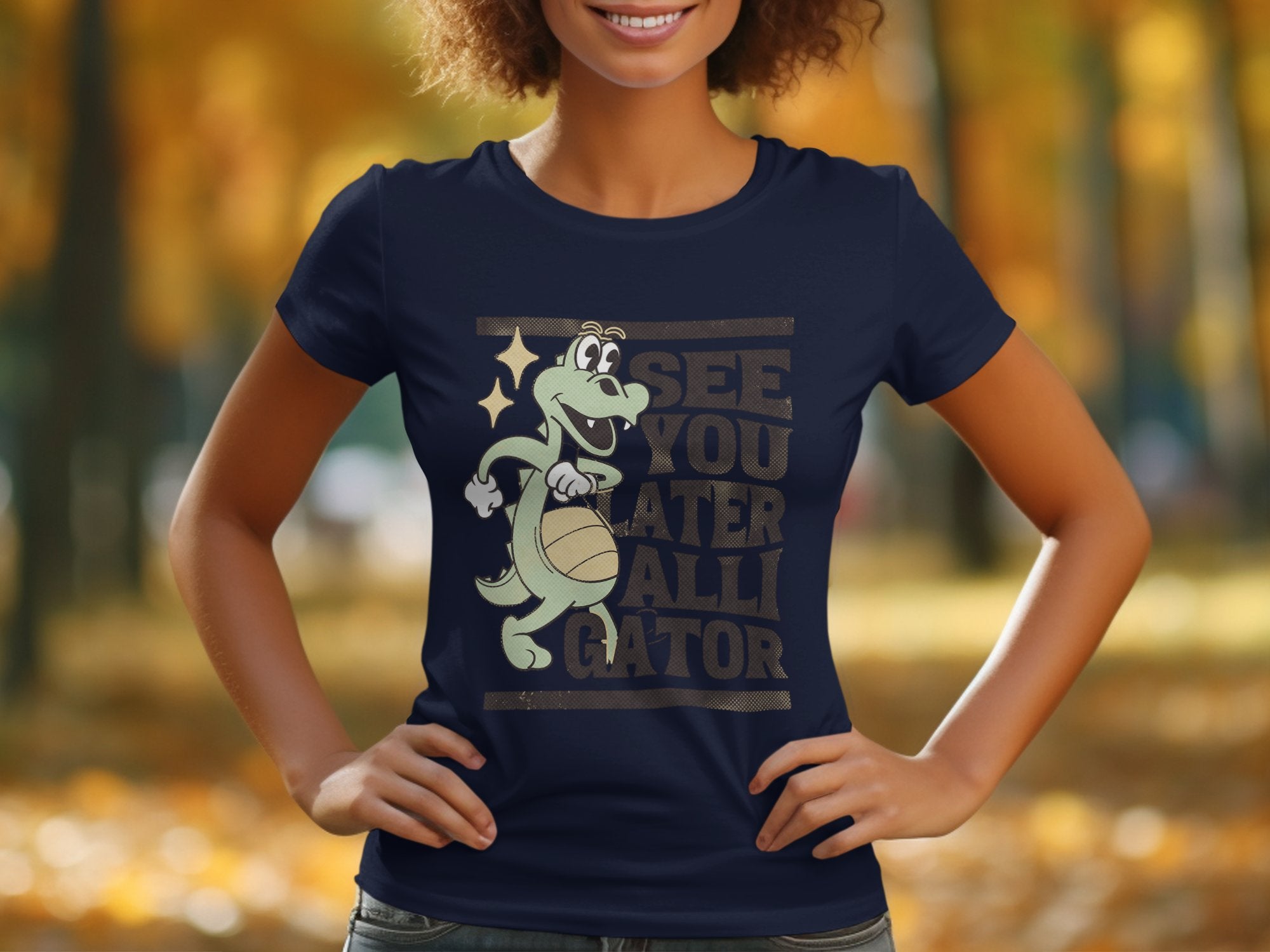 A woman wears a navy blue Garment Graphics Alligator T-Shirt, showcasing a cartoon alligator with a basketball. The shirt humorously reads, See you later alligator, set against blurred autumn trees with vibrant yellow leaves.