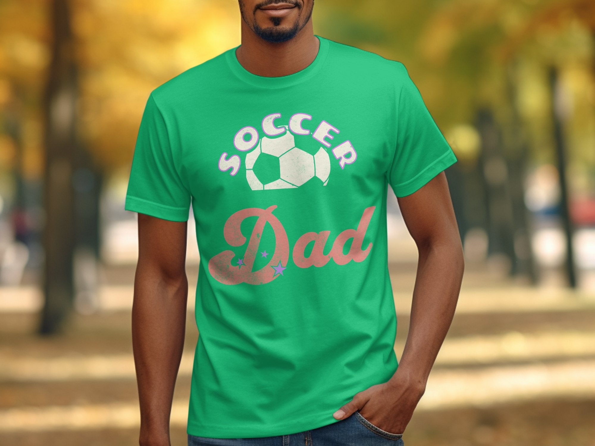 A person is outdoors in a green Soccer Dad graphic T-shirt from Garment Graphics with a soccer ball design. The backdrop features a blurred pathway flanked by trees in bright autumn colors, enhancing the scenes charm.