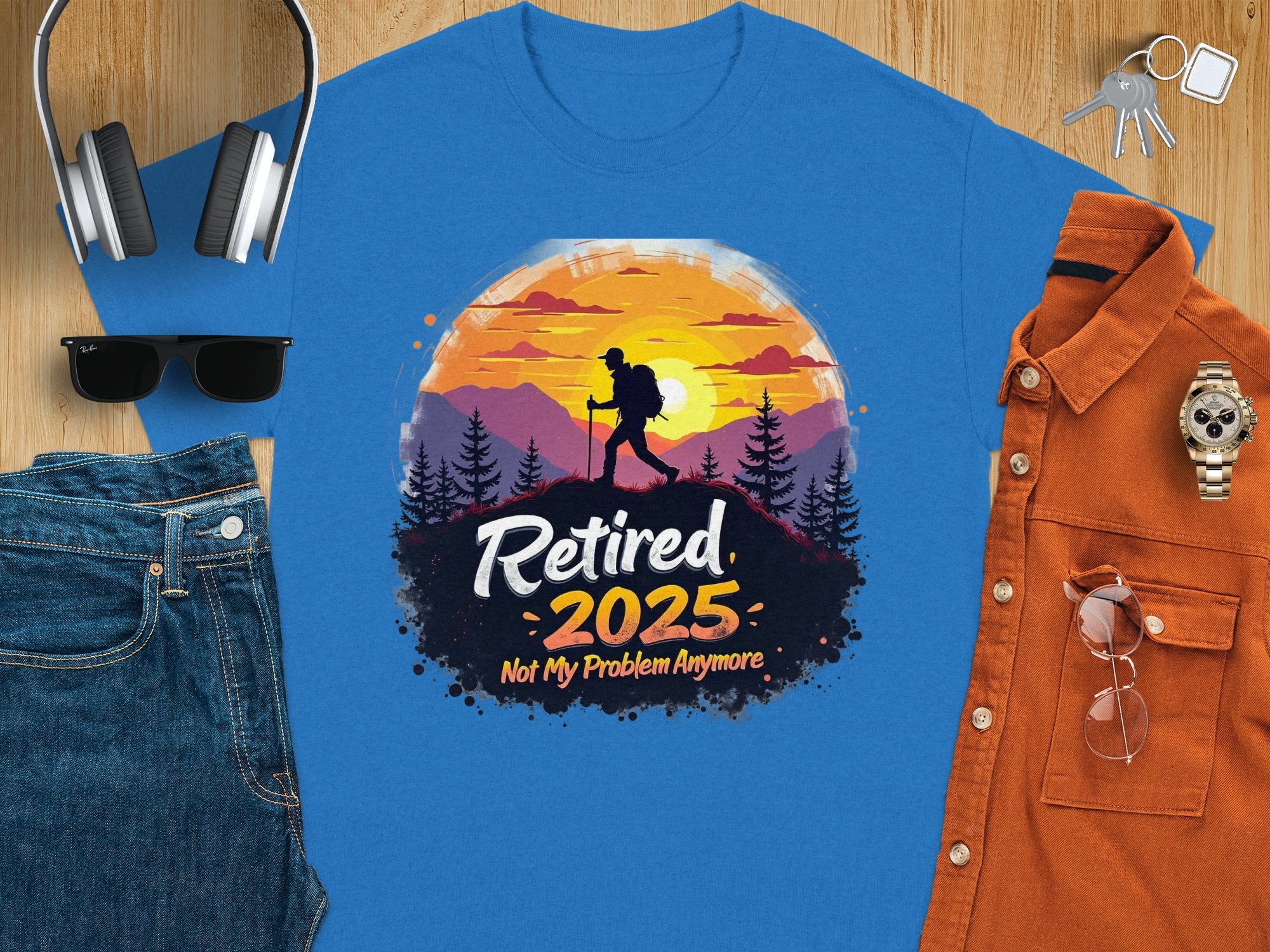 A flat lay features Garment Graphics blue T-shirt with a hiking silhouette and Retired 2025, Not My Problem Anymore text, celebrating retirement. Its paired with headphones, sunglasses, jeans, keys, a watch, and an orange shirt on a wood surface.