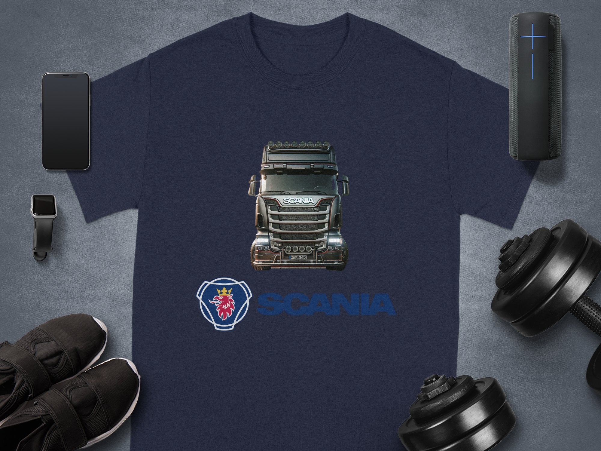 Garment Graphics offers a stylish navy blue T-shirt featuring a Scania truck and logo. Ideal for truck fans, its paired with modern essentials: smartphone, smartwatch, black sneakers, portable speaker, and dumbbell.