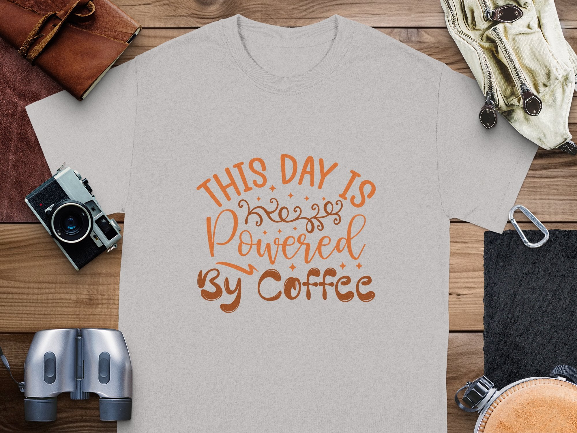 A classic fit, grey Garment Graphics t-shirt boasts This day is powered by coffee in bold orange and brown. Set on a wooden surface, its paired with a camera, binoculars, and a leather wallet, all evoking adventure. Ideal for coffee-loving t-shirt fans!.