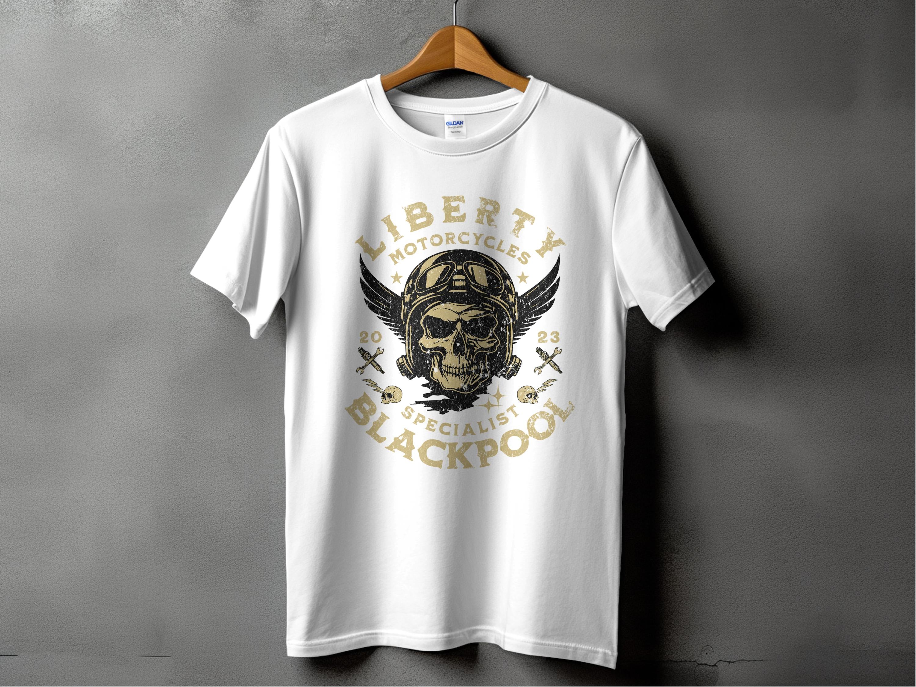 A white T-shirt by Garment Graphics on a wooden hanger against a gray backdrop, ideal for motorcycle fans. It features a vintage skull with goggles and wings, encircled by Liberty Motorcycles Specialist Blackpool 2023 and skull-and-crossbones icons.