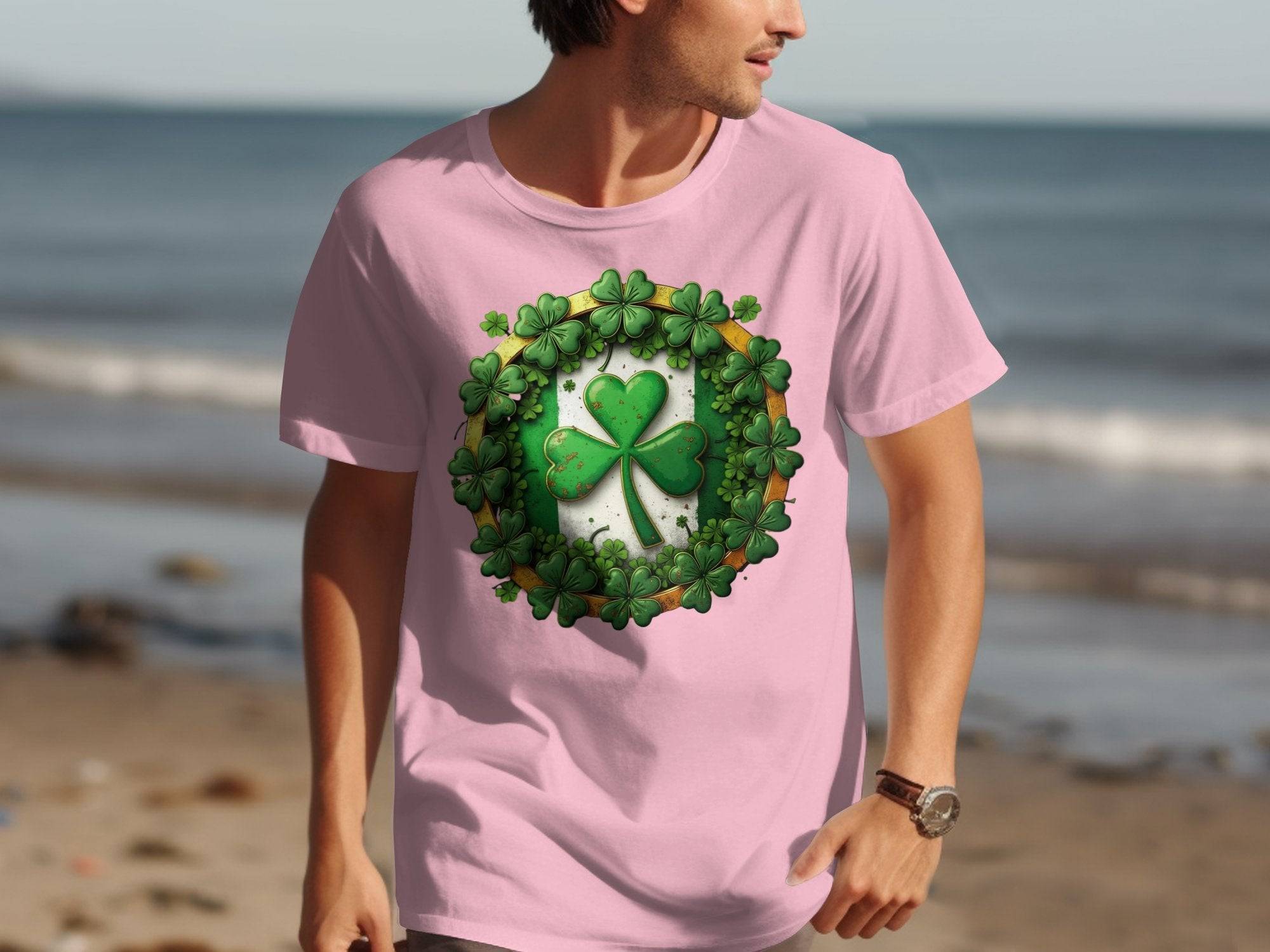 A man walks along the beach in a vibrant pink T-shirt from Garment Graphics, ideal for St. Patricks Day. It showcases a large green shamrock encircled by smaller ones in a ring pattern, against the scenic ocean waves and sandy shore backdrop.