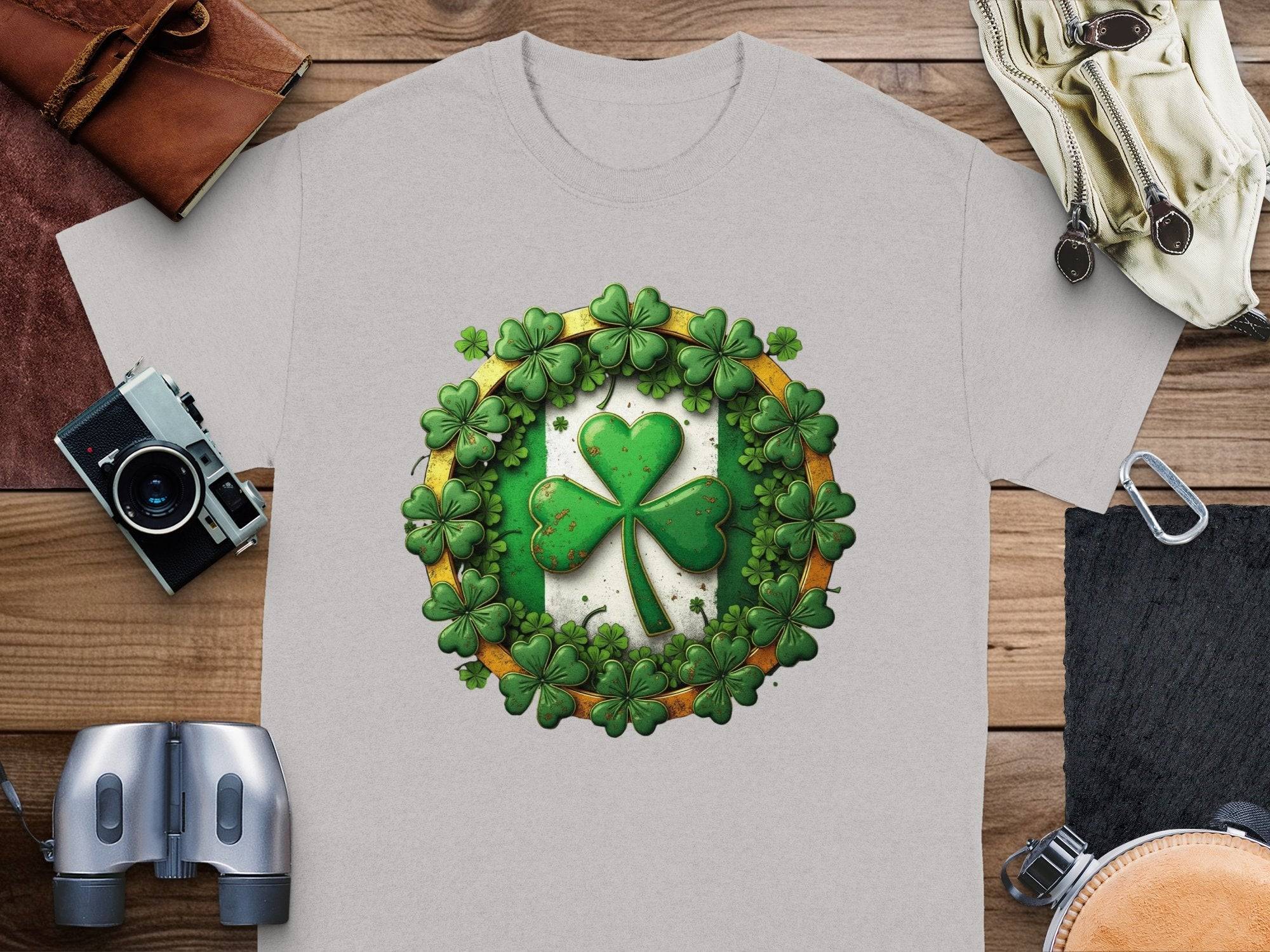 A light gray Garment Graphics T-shirt featuring a green shamrock clover framed by shamrock leaves rests on a wooden surface, ideal for St Patricks Day. Nearby are items like a vintage camera, leather pouch, and binoculars.
