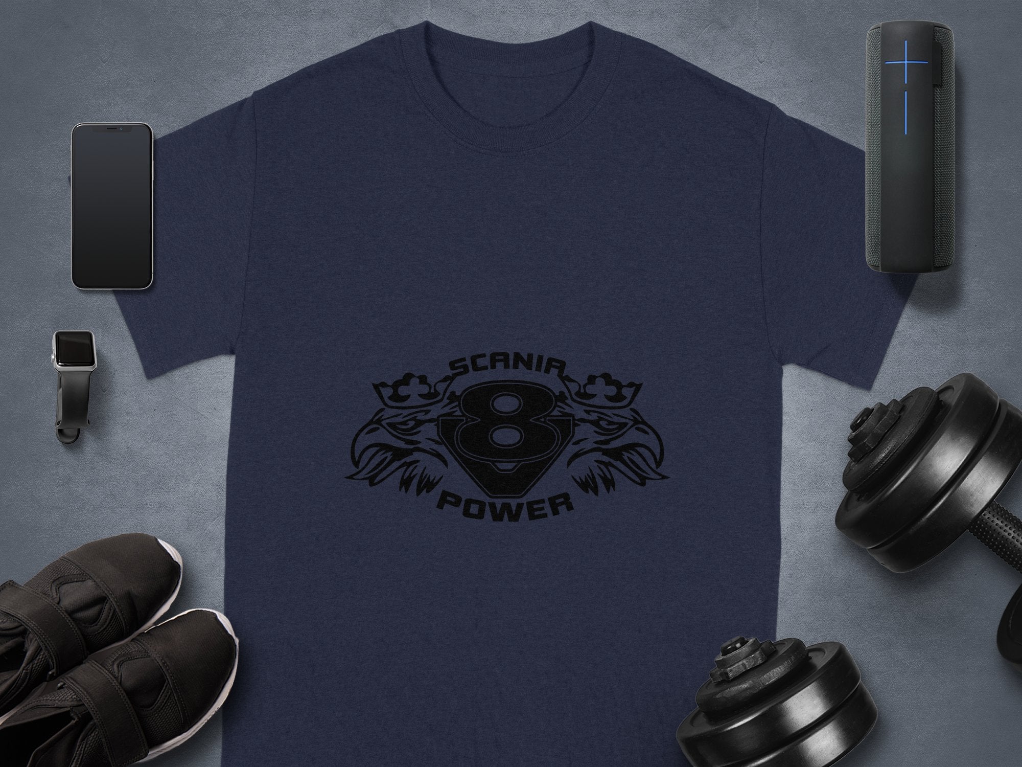 Flat lay featuring a Garment Graphics navy blue T-shirt with Scania Power Eagle and V8 engine design, alongside a smartphone, smart speaker, smartwatch, dumbbells, and black sneakers on a gray surface, highlighting the synergy between tech accessories and graphic design fashion.