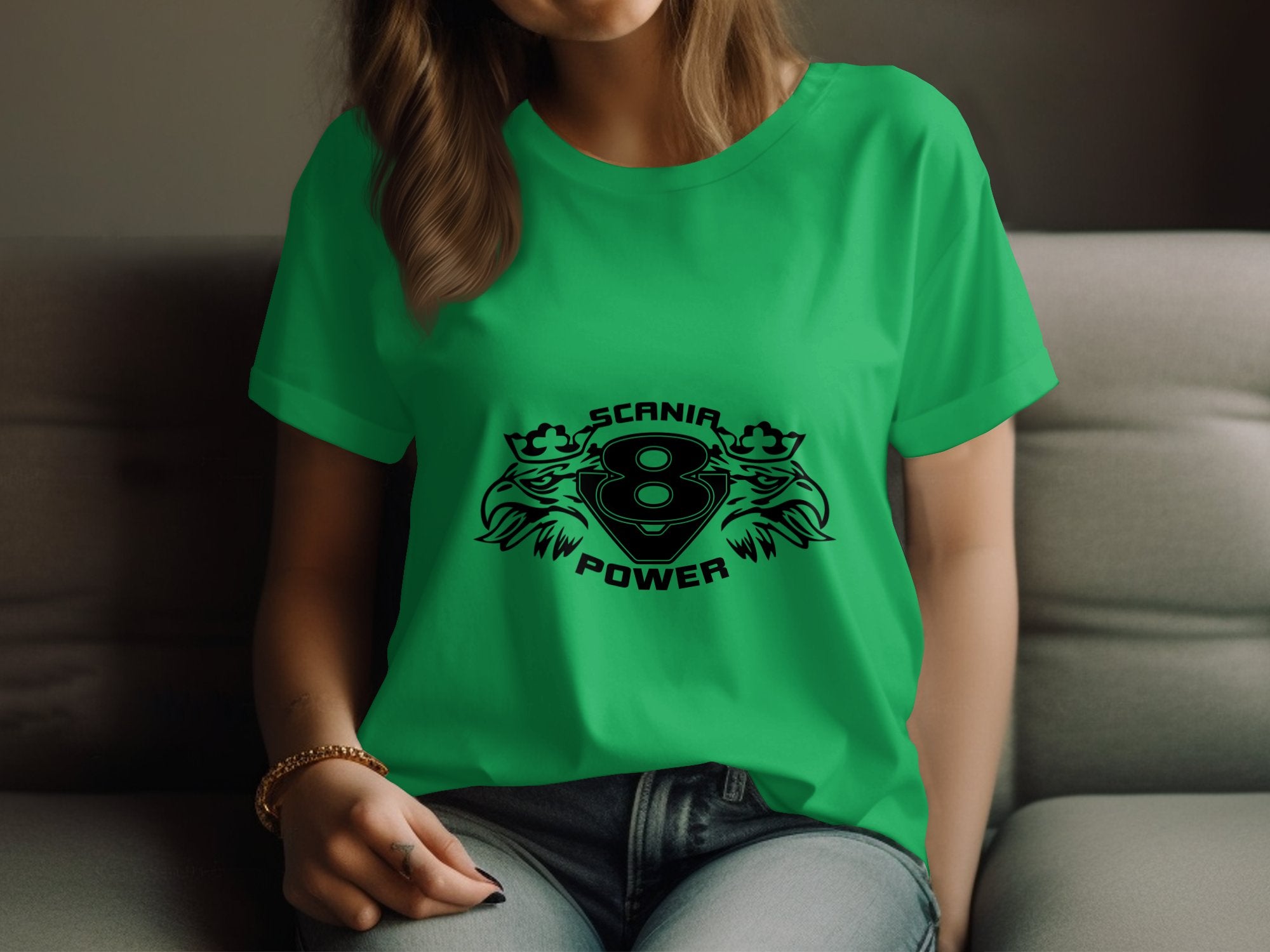 A person relaxes on a sofa, wearing a bright green Garment Graphics T-shirt with a black design featuring the number 8 encircled by crowned lions and the words SCANIA POWER, flanked by stylized eagle heads. Theyre also wearing jeans and a bracelet.