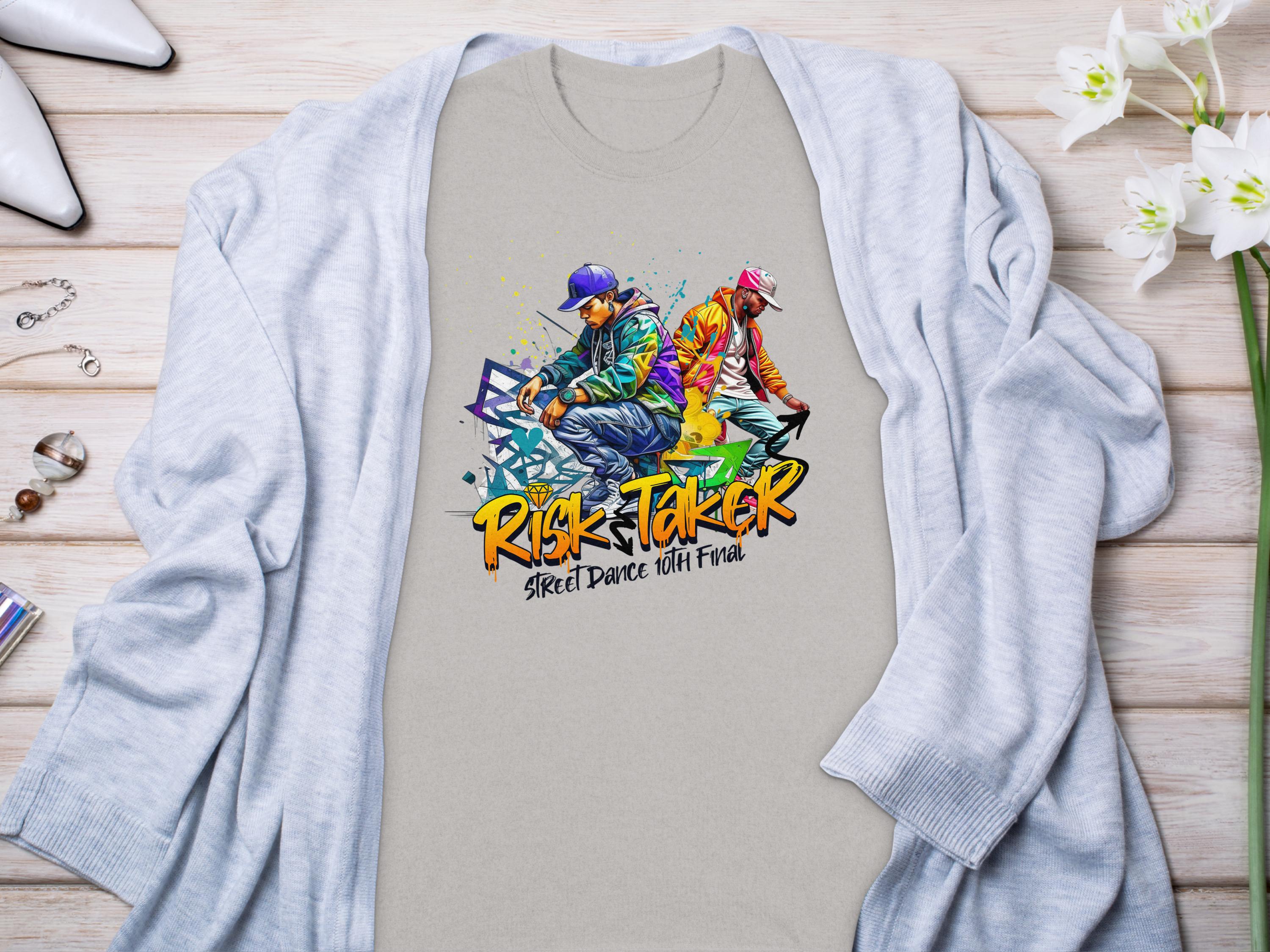 A vibrant light gray t-shirt from Garment Graphics features a street dance graphic of two dancers and the text Risk Taker above Street Dance with Fran, celebrating the Risk Taker 10th Final T-Shirts. Displayed on wood, it pairs with a blue cardigan, accessories, and white flowers.
