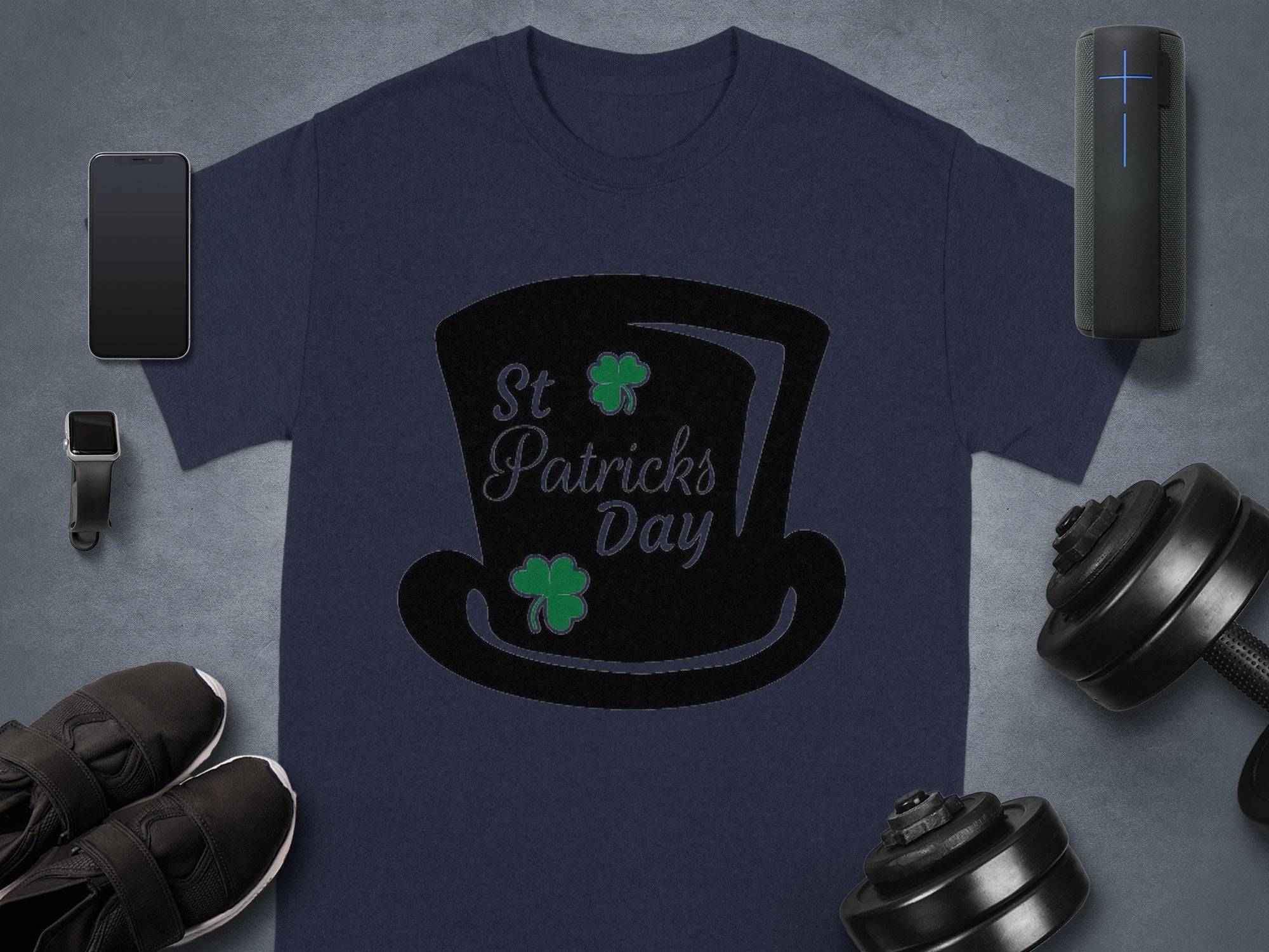 Celebrate in style with Garment Graphics navy blue T-shirt adorned with a black top hat, two shamrocks, and St Patricks Day text. A smartphone, smartwatch, shoes, dumbbells, and a portable speaker create a festive scene against a gray background.