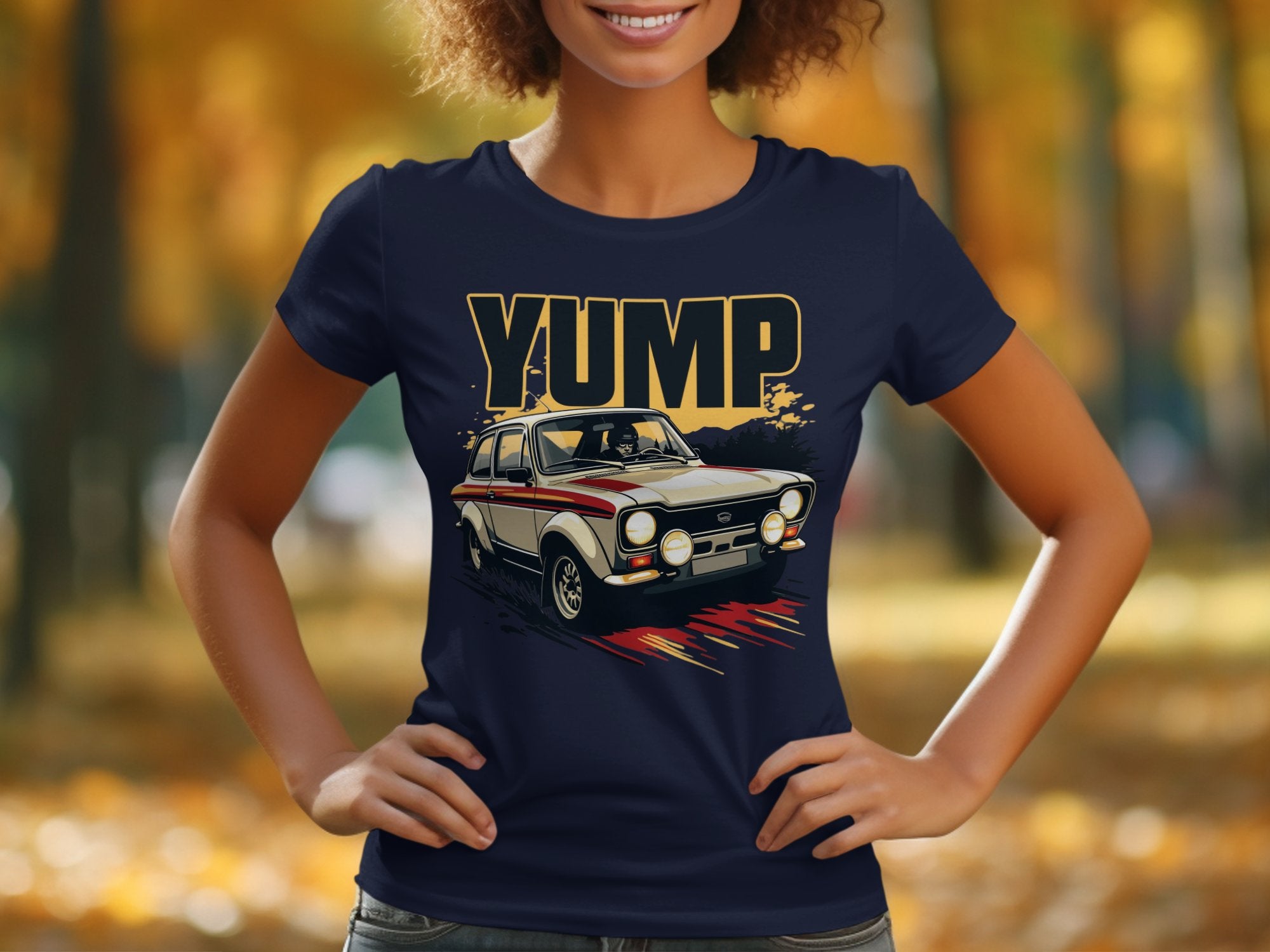 A person stands with hands on hips, wearing a dark blue retro Garment Graphics t-shirt featuring a vintage car jumping, with the bold graphic YUMP above it. The background is an autumn scene with blurred trees and yellow leaves.