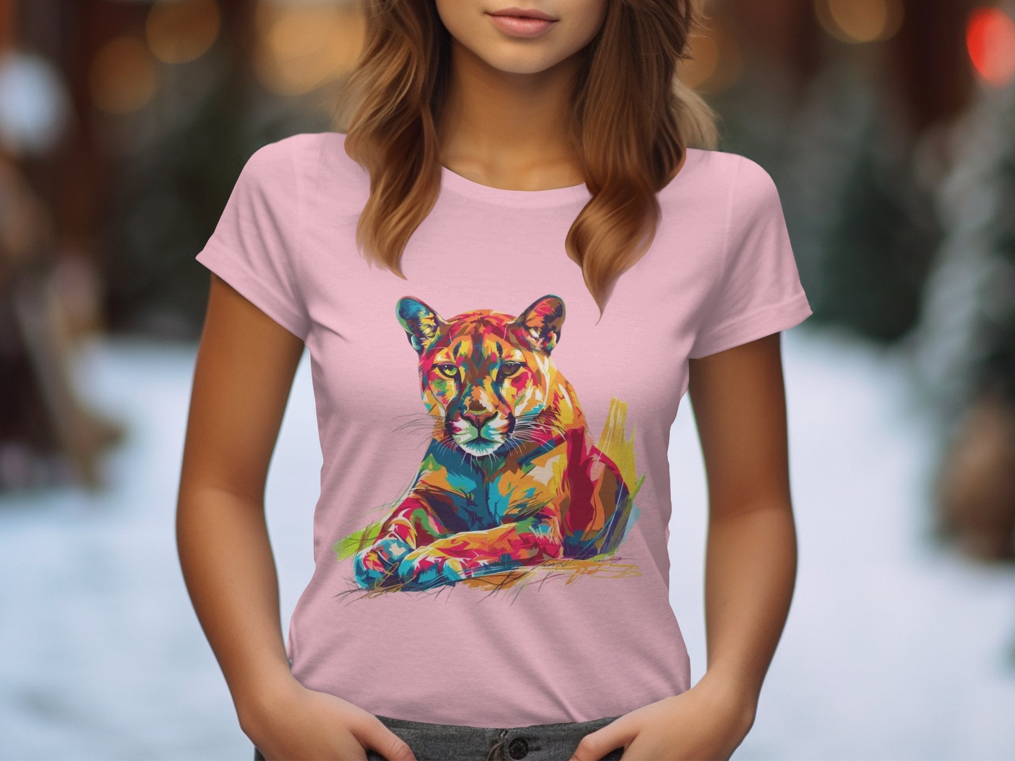 A woman wearing a Garment Graphics pink T-shirt with a vivid resting cougar graphic print stands against a softly blurred, snowy background illuminated by warm lighting, concealing her face.