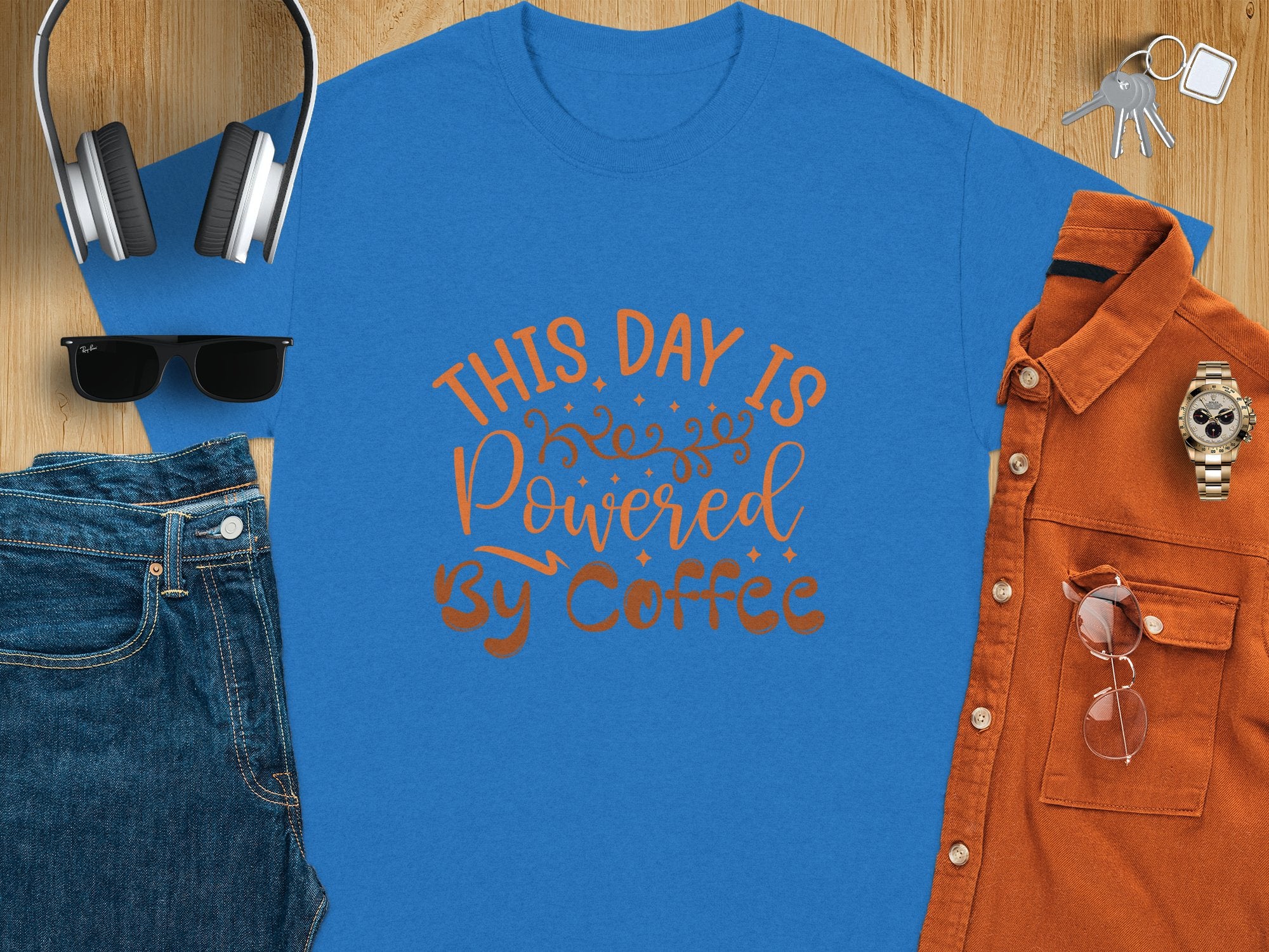 A blue classic fit T-shirt from Garment Graphics, featuring THIS DAY IS Powered BY Coffee, lies flat on a wooden surface alongside jeans, sunglasses, headphones, a brown shirt, and a watch, exuding positivity.