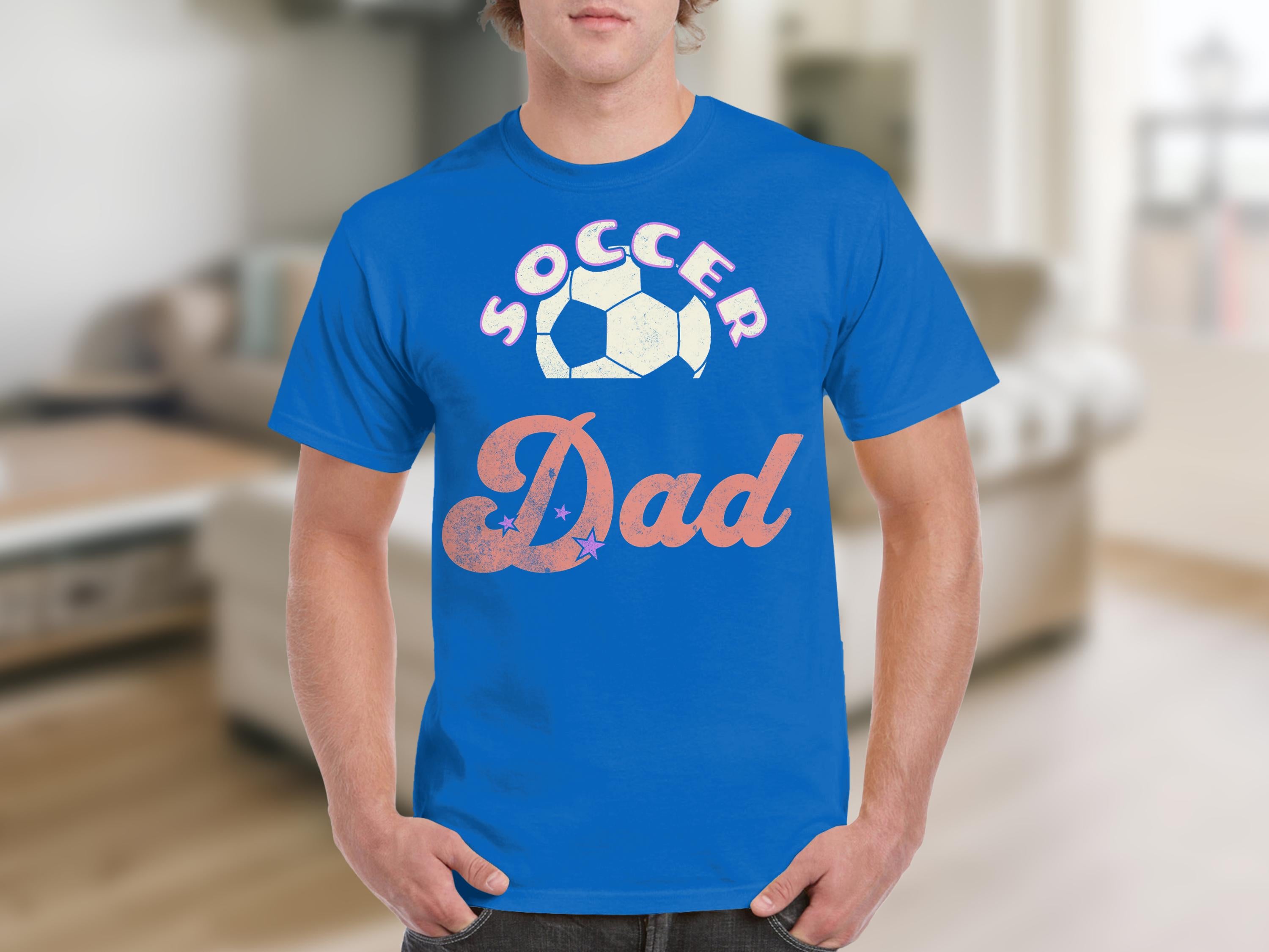 A person indoors wears Garment Graphics blue Soccer Dad T-shirt, where the o is a soccer ball design. The relaxed vibe of the blurred living room contrasts with the shirts bold statement.