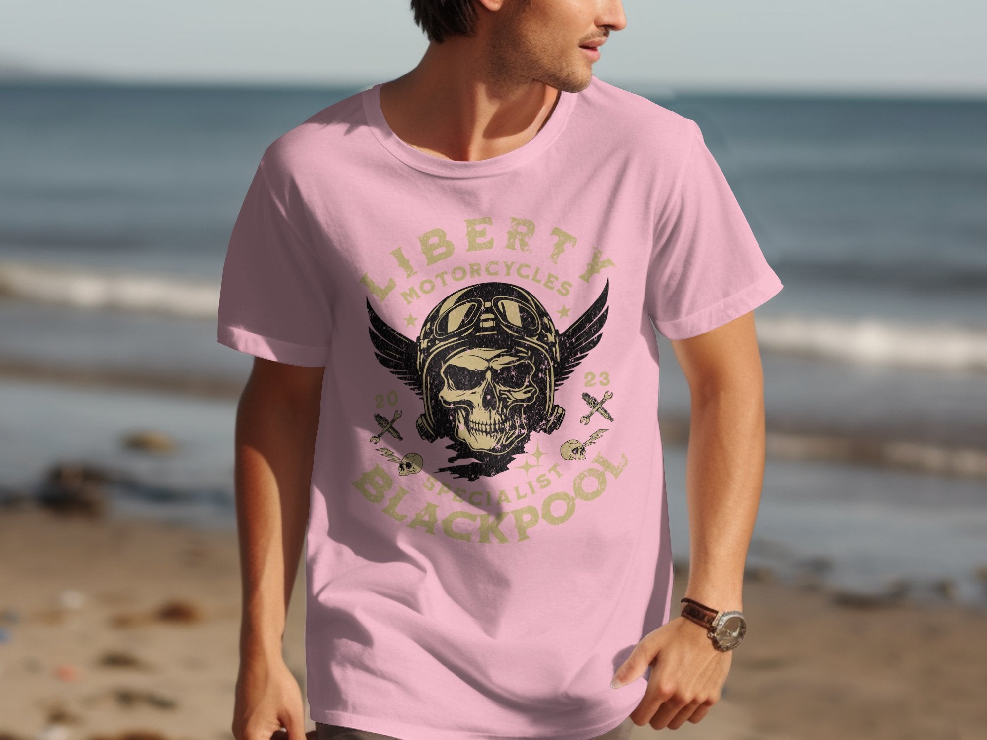 Sporting a pink t-shirt from Garment Graphics, featuring a vintage skull and wings design with Liberty Motorcycles Steel Artist Blackpool 2023, someone strolls along the beach, ocean in view—a must for motorcycle fans embracing seaside freedom.