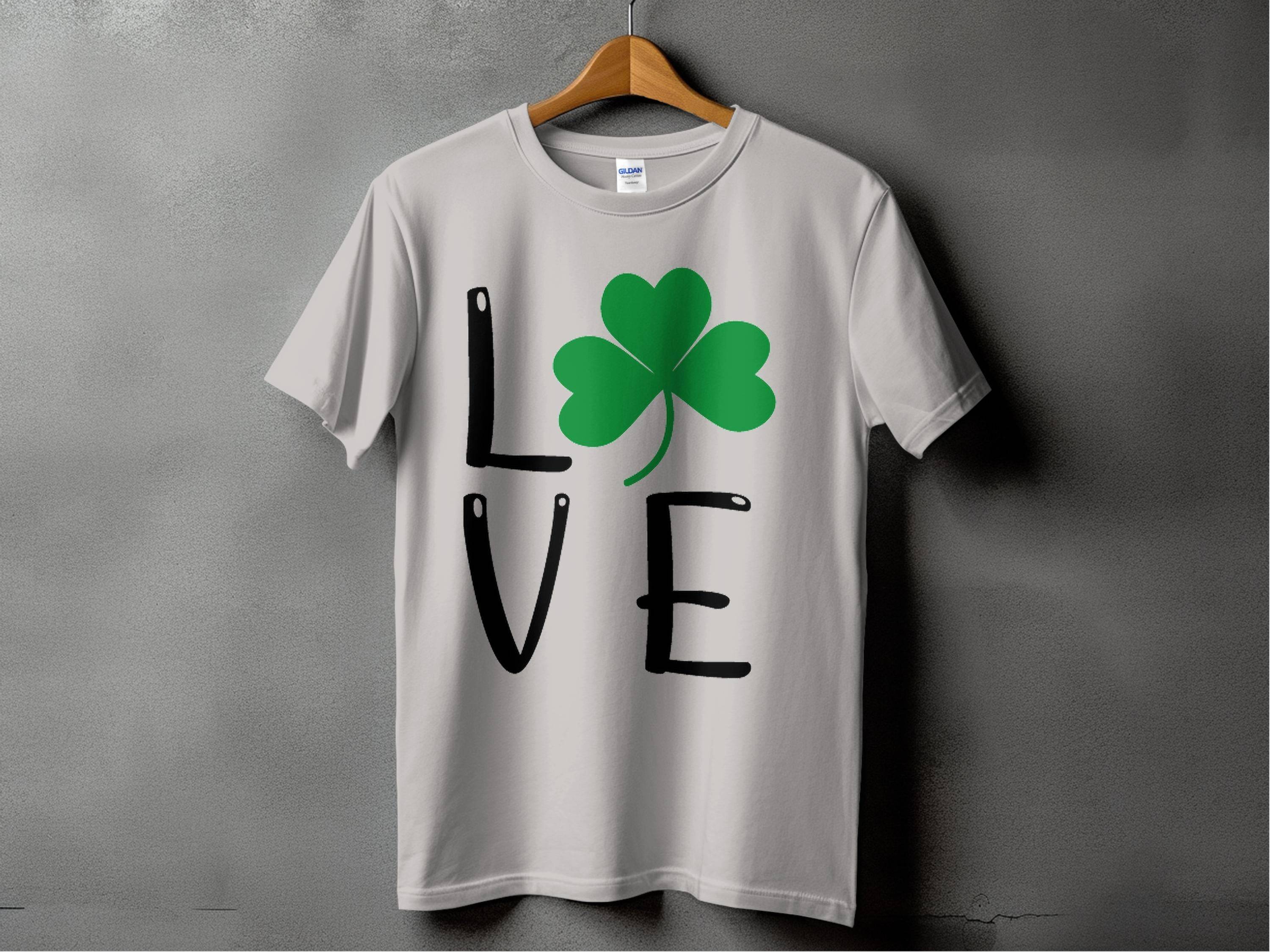 This Sport Grey T-shirt on a hanger is, featuring L♥VE with a shamrock replacing the O, embodies Irish spirit against a plain gray wall for St. Patricks Day.