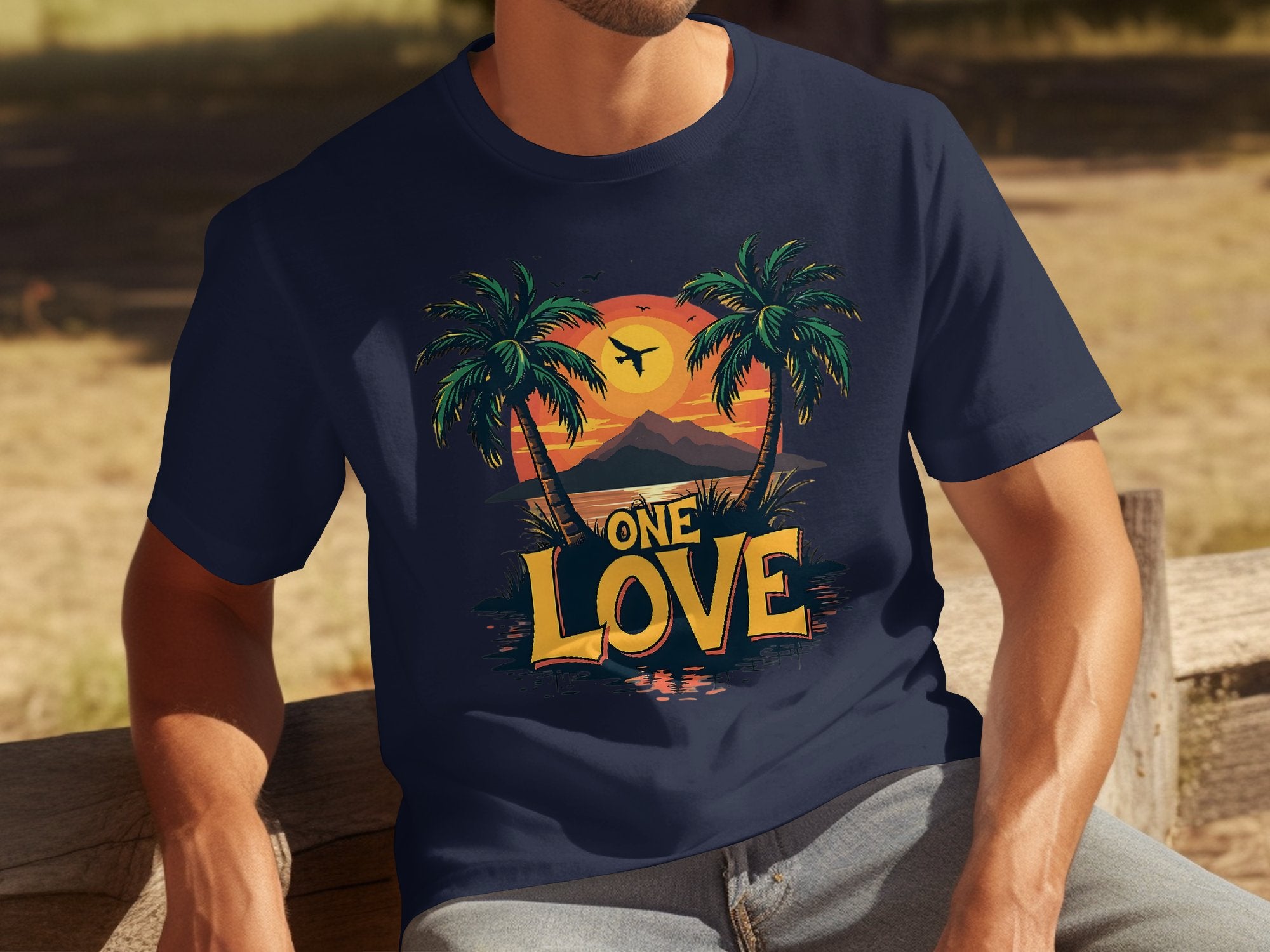 Someone wears a Garment Graphics navy blue t-shirt featuring a lively graphic of two palm trees, a tropical sunset, and an airplane, with ONE LOVE below. A blurred natural setting in the background enhances this ode to beach lovers.