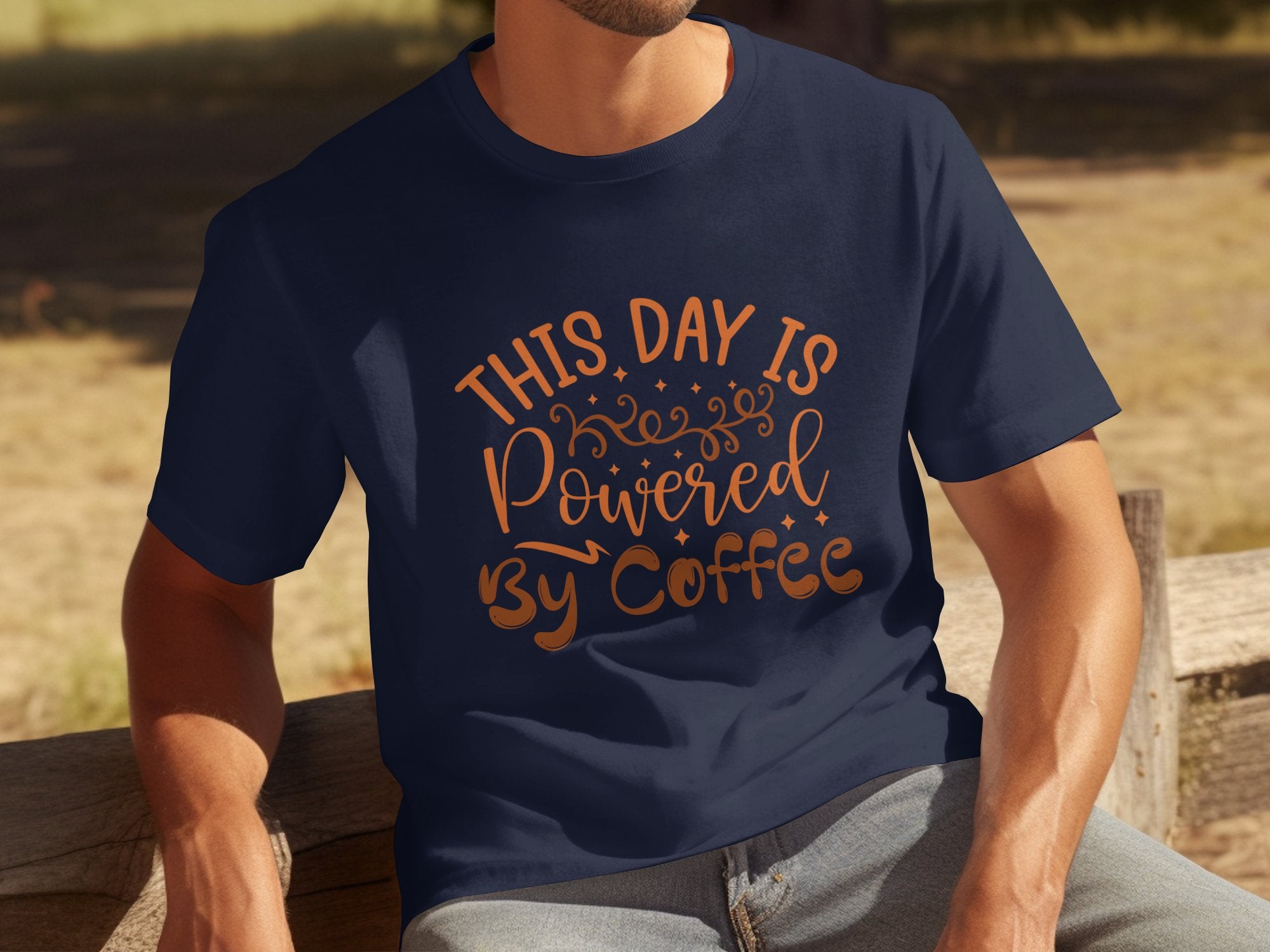 Wearing a classic fit navy blue tee from Garment Graphics, emblazoned with This Day Is Powered By Coffee, someone sits on a wooden bench, radiating positivity in an inviting outdoor setting.