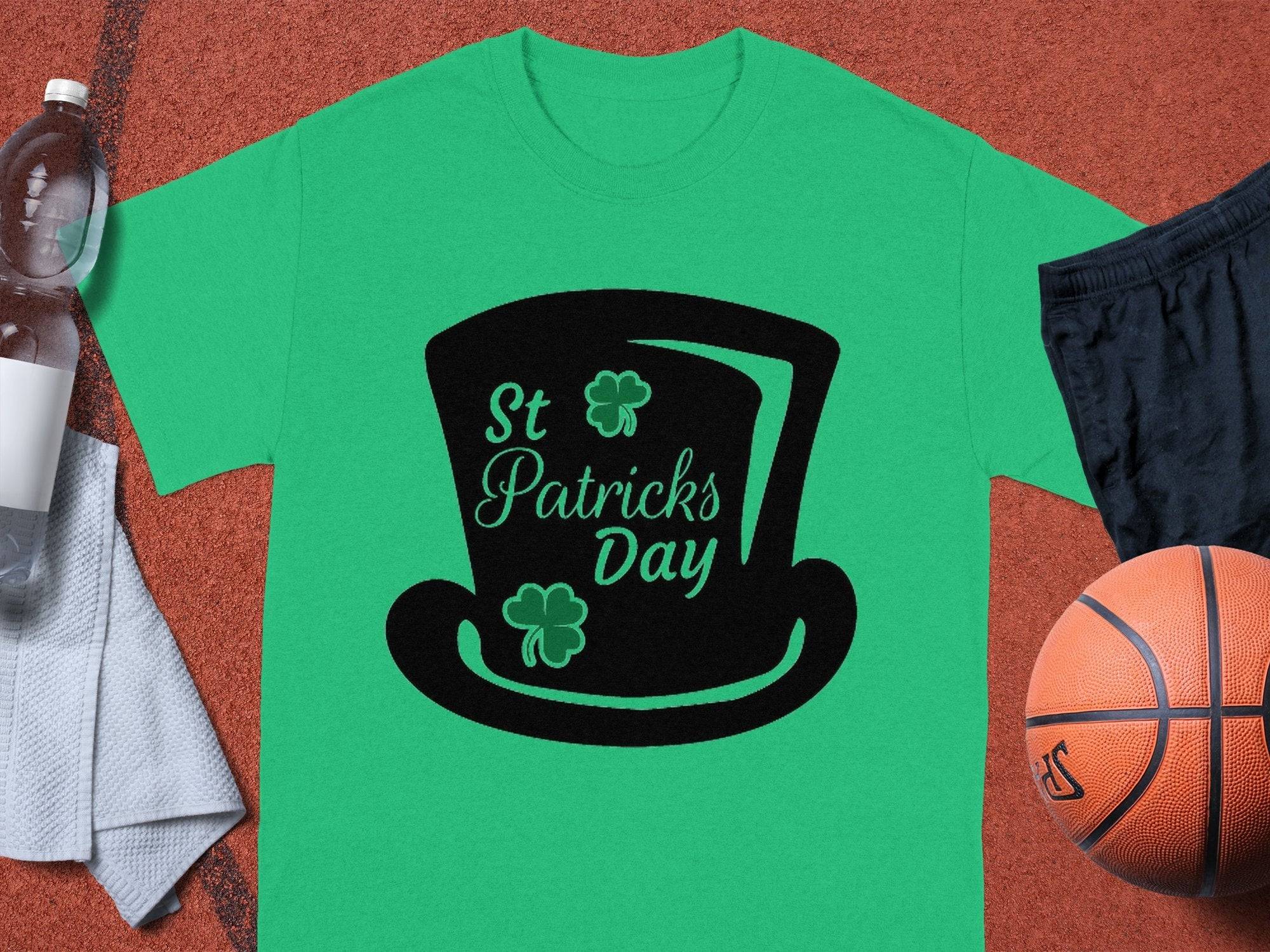 A Garment Graphics green T-shirt with a black top hat graphic, St Patricks Day text, and shamrocks is set on a sports court. A basketball, black shorts, and water bottle nearby perfectly capture the playful spirit of St Patricks Day.
