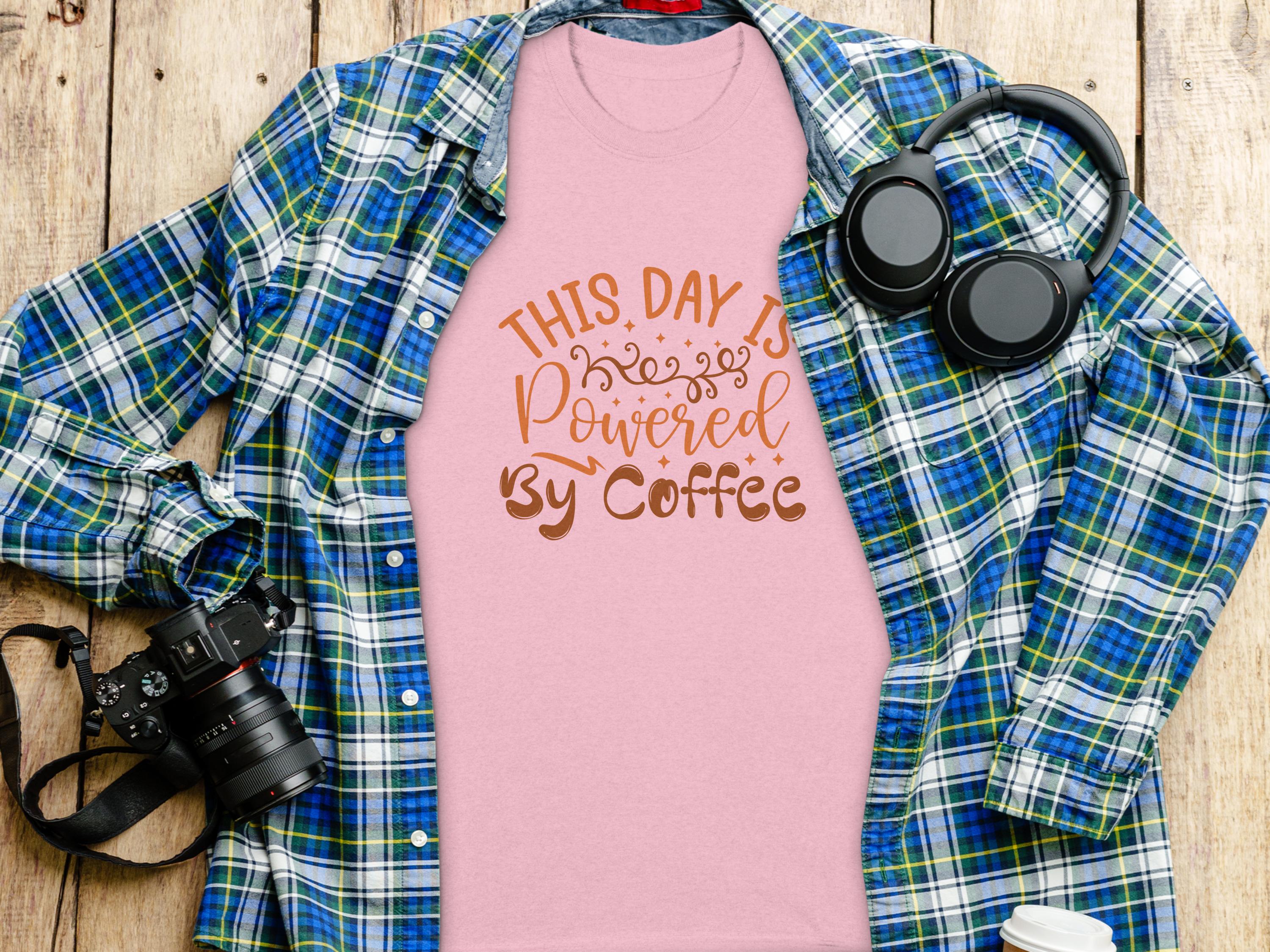 A pink T-shirt from Garment Graphics with This Day is Powered by Coffee sits on a wooden surface, radiating positivity. Its paired with a green and blue plaid shirt for layering, alongside headphones, a camera, and a disposable coffee cup.