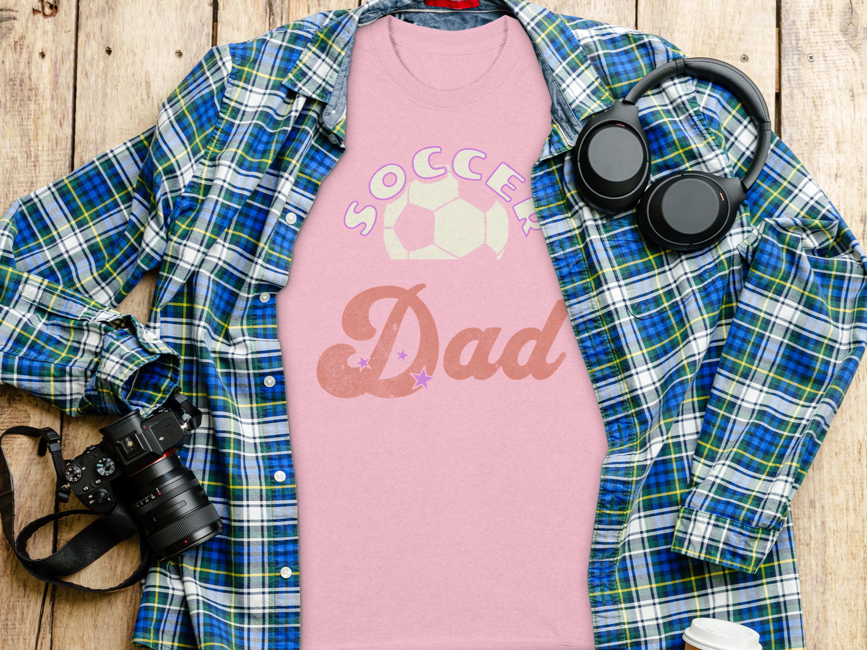 A pink Soccer Dad T-shirt from Garment Graphics is styled with a blue and green plaid shirt, alongside black headphones, a camera, a coffee cup lid, and a phone on a wooden surface.