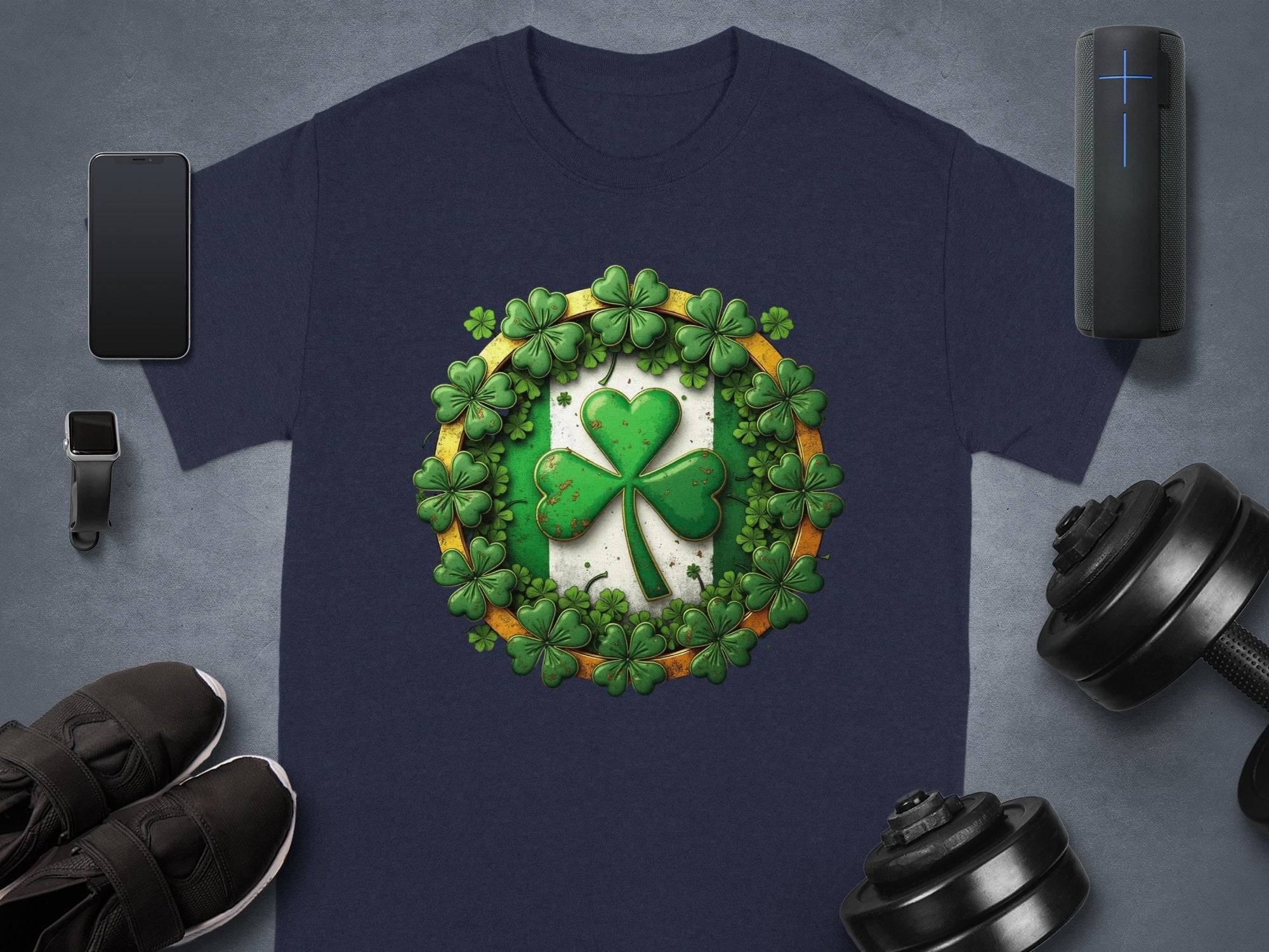 A Garment Graphics navy blue St. Patricks Day T-shirt with a shamrock and clover design is neatly laid on a gray surface, surrounded by essentials like a smartphone, smartwatch, Bluetooth speaker, shoes, and dumbbells.