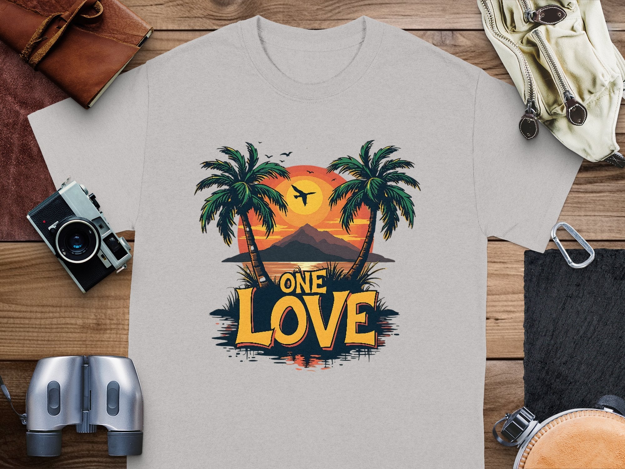 Garment Graphics offers a light gray T-shirt ideal for beach lovers, showcasing a tropical sunset print with palm trees, a plane, and “ONE LOVE” boldly displayed. Styled on wood and paired with essentials like a camera, binoculars, wallet, and small bag.