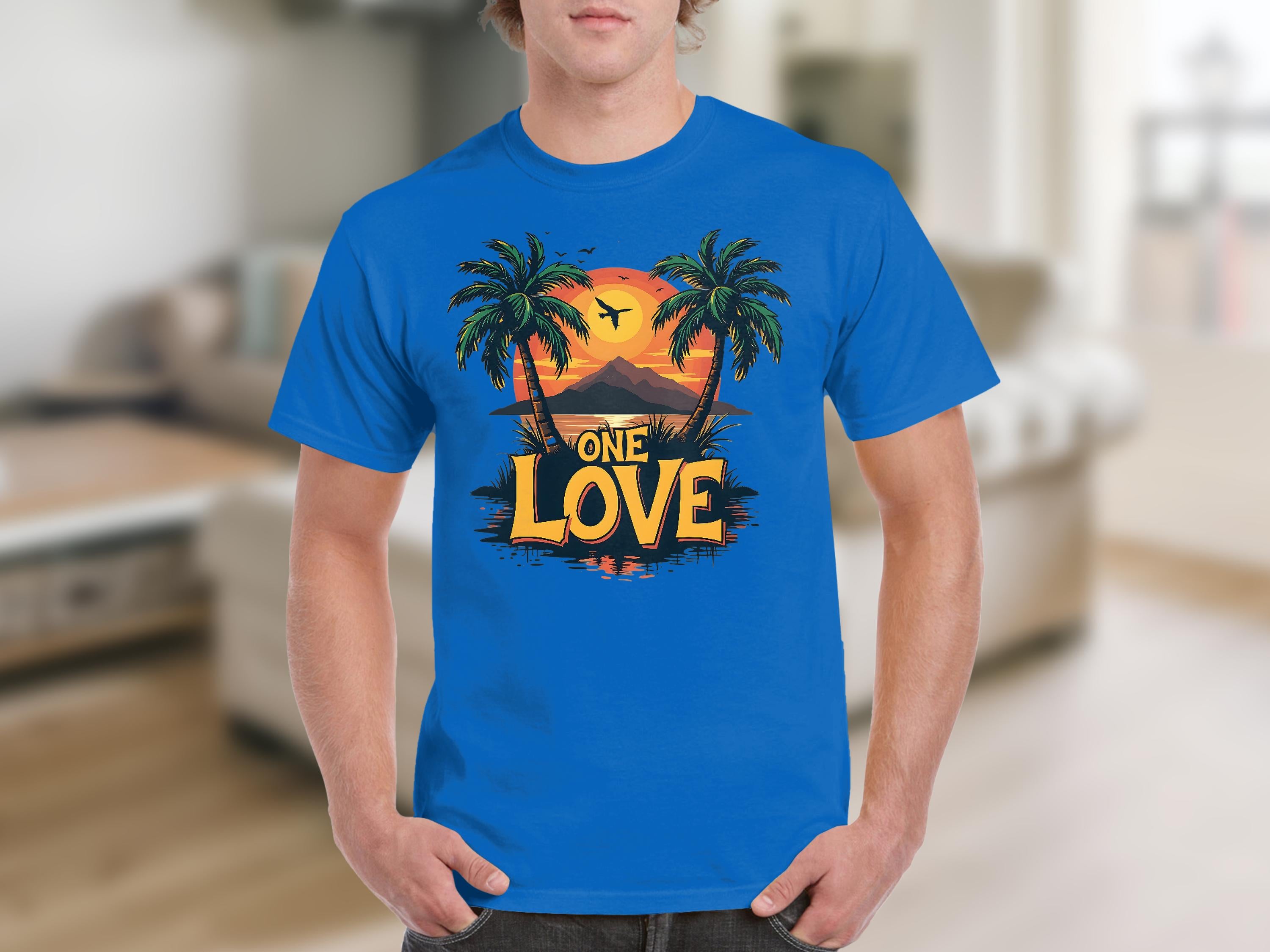A man wears a blue Garment Graphics t-shirt showing a tropical sunset with palm trees and an airplane. Ideal for beach lovers, it features One Love in a stylized font. The indoor setting has neutral-toned furniture and blurred details.