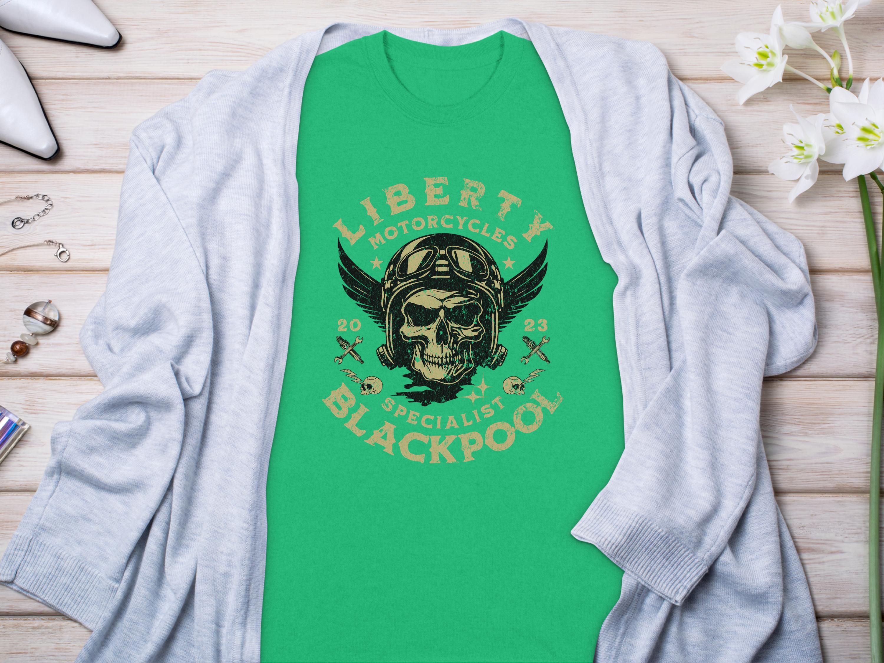 A green Garment Graphics t-shirt with a skull, helmet, and wings design labeled Liberty Motorcycles Blackpool 2023 lies on light wood. Perfect for motorcycle enthusiasts, its surrounded by a gray cardigan and accessories.