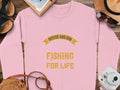 A pink sweatshirt from Garment Graphics showcases the text Father and Son Fishing for Life in bright yellow, styled with a hat, sunglasses, notebook, instant camera, and sandals on a wooden surface.