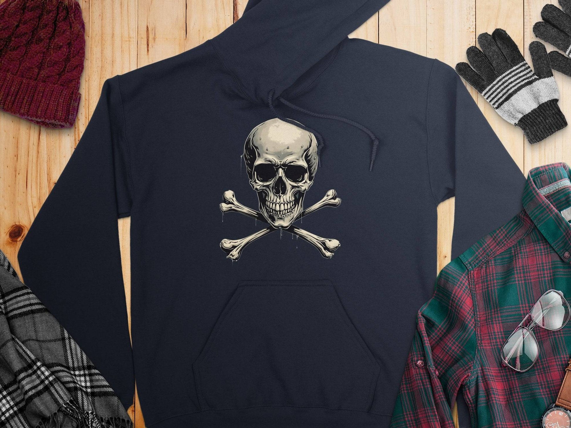 A Garment Graphics black hoodie featuring a trendy skull and crossbones design is displayed on wood, surrounded by a red knit hat, striped gloves, a gray plaid scarf, and a green and red plaid shirt.