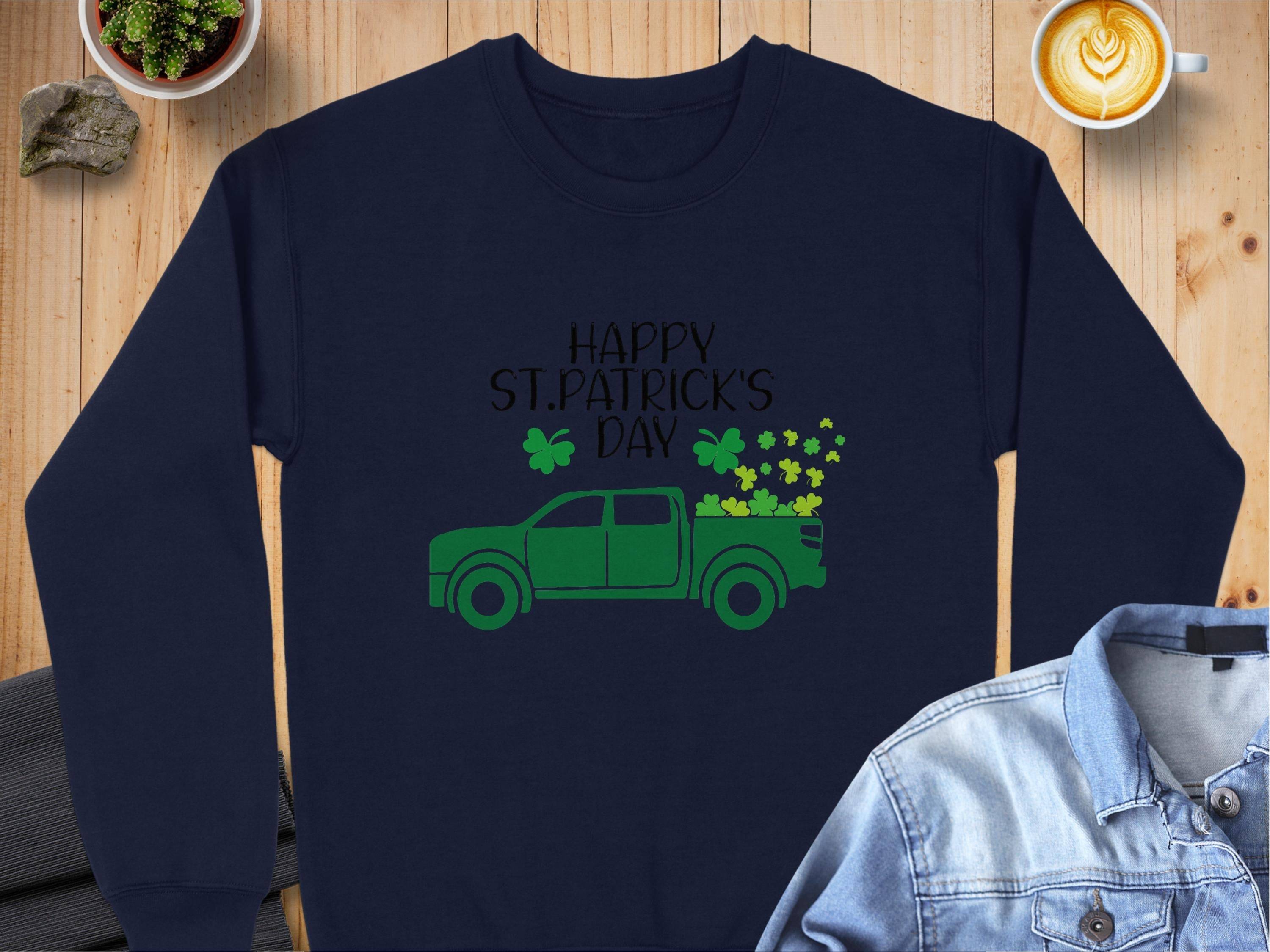 A festive navy blue sweatshirt from Garment Graphics features a green truck with shamrocks and Happy St. Patricks Day on a wooden surface, complemented by a denim jacket, heart-art latte, and small plant for a vibrant scene.