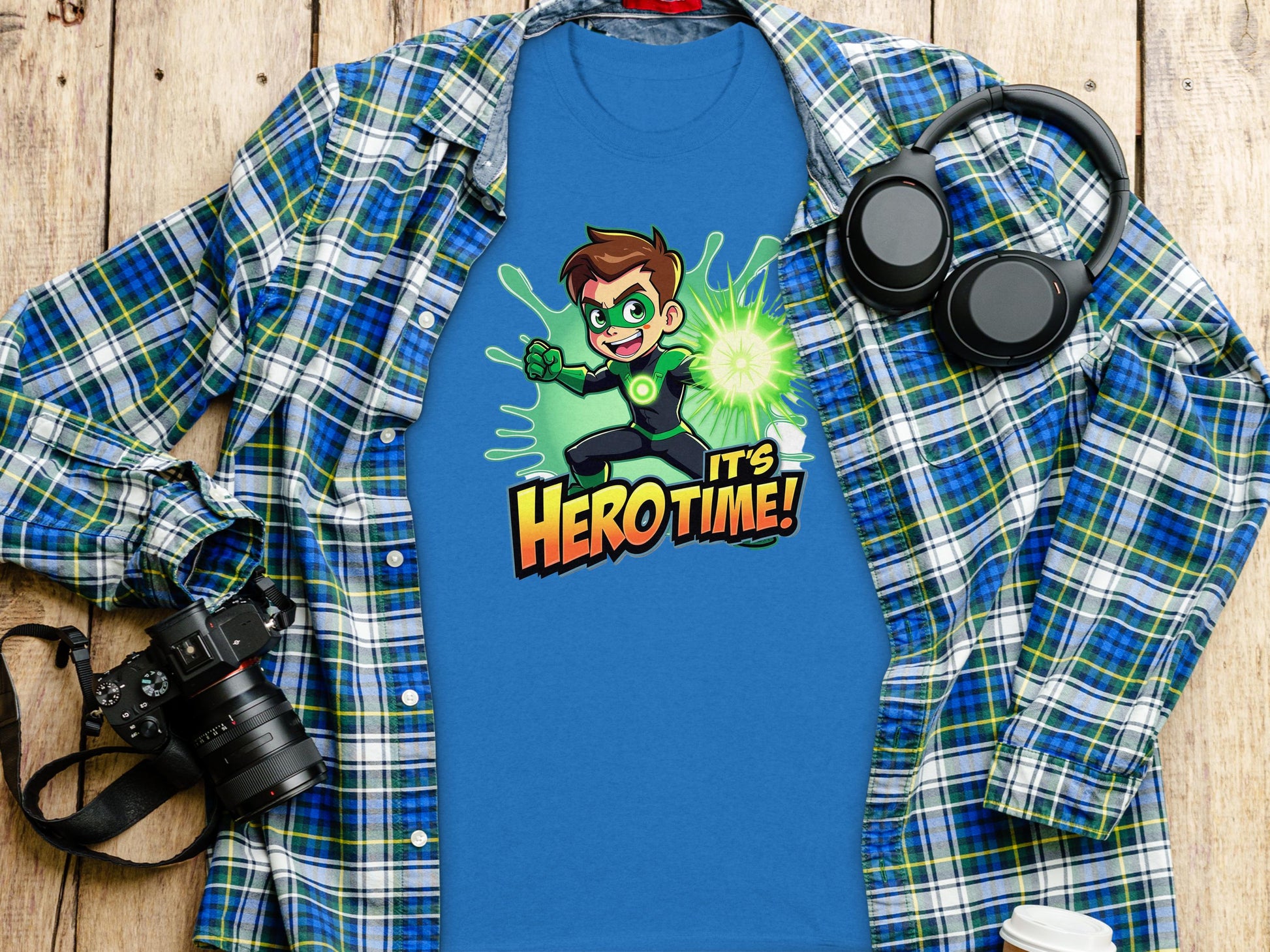 Displayed on a wooden surface, the Garment Graphics blue T-shirt boasts a superhero graphic with Its Hero Time! text, paired with a blue and green plaid shirt, a camera, headphones, and a white cup. Ideal for fashionable family sizing.