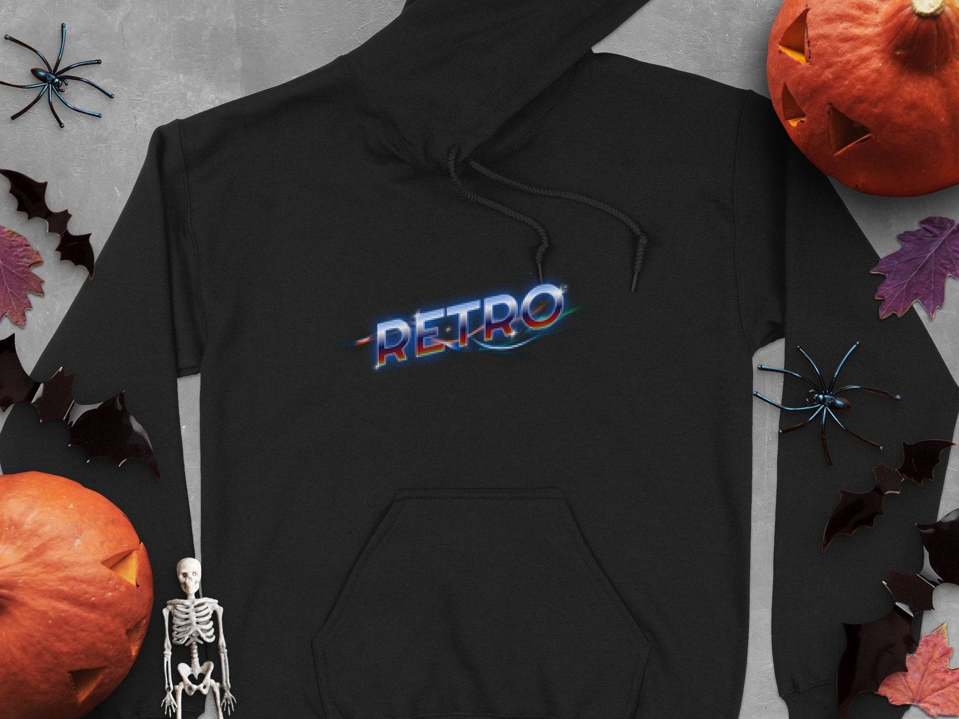 A black Garment Graphics hoodie featuring RETRO in vivid glitch-style text stands out, creating a trendy vibe. Its surrounded by Halloween-themed items like pumpkins, a skeleton figure, bats, and spiders against dark autumn leaves on gray.