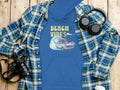 A Garment Graphics hoodie featuring a Beach Vibes vintage car and wave graphic sits under a blue-green plaid shirt. Nearby on the wooden surface are black headphones, a camera, and a white cup, embodying retro beach style.