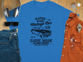 A flat lay of a blue T-shirt with Vintage Car Club graphics from Garment Graphics, headphones, sunglasses, jeans, a rust-colored button-up shirt, and a wristwatch on a wooden surface—ideal for Retro Automotive enthusiasts.
