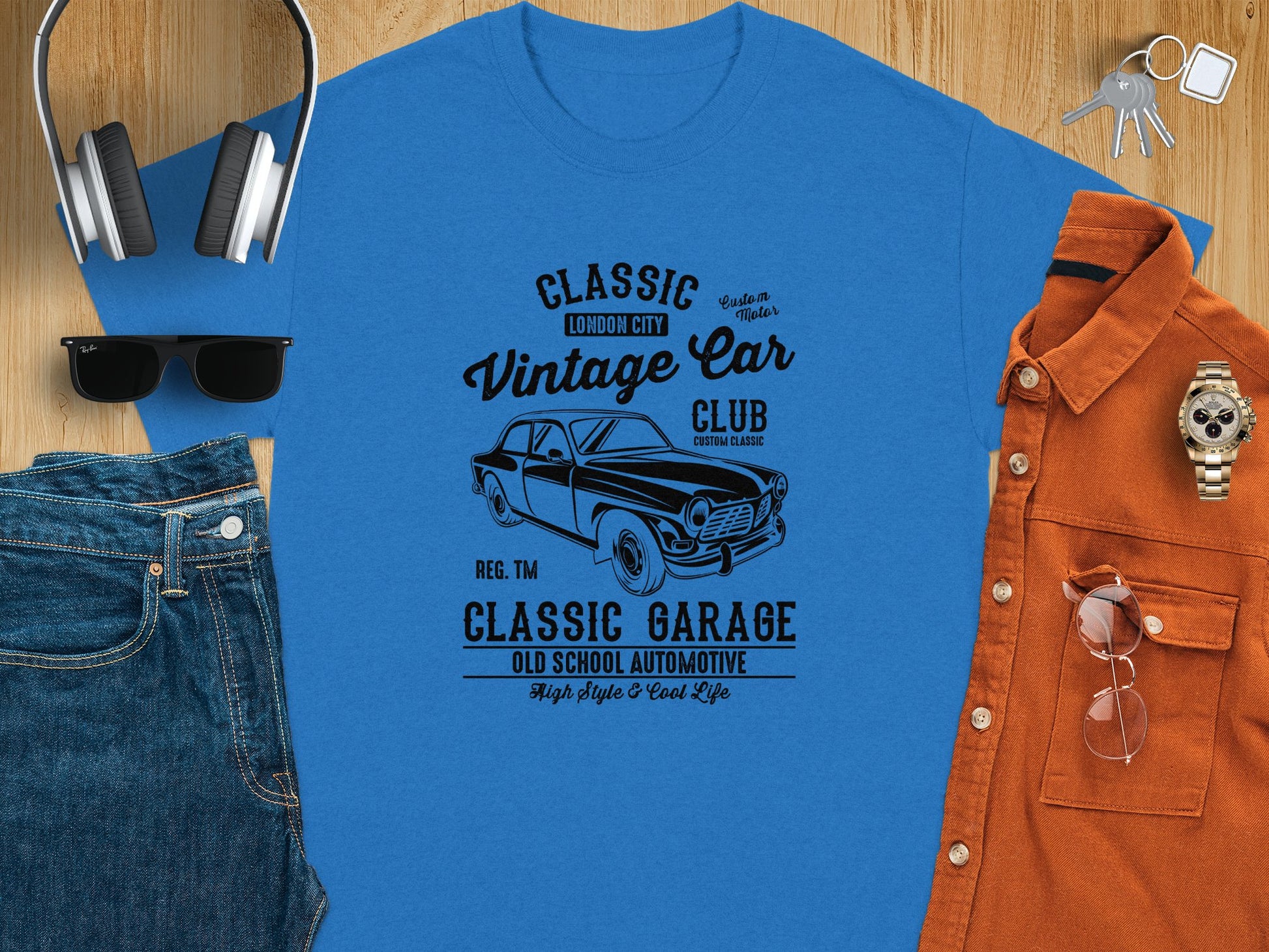 A flat lay of a blue T-shirt with Vintage Car Club graphics from Garment Graphics, headphones, sunglasses, jeans, a rust-colored button-up shirt, and a wristwatch on a wooden surface—ideal for Retro Automotive enthusiasts.