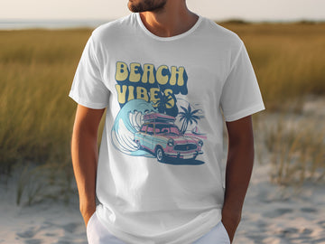 A person wears a white Garment Graphics t-shirt featuring a classic car with surfboards, palm trees, and waves. The vibrant design highlights the words Beach Vibes. Sand and grass provide an ideal backdrop for this summery scene.