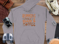 Garment Graphics classic fit gray hoodie, featuring Running on Grace & Coffee in vibrant orange and white lettering. Made from medium-heavy fabric, its ideal for adventurers. Displayed with a camera, binoculars, leather-bound book, bag, gloves, and carabiner on a wooden surface.