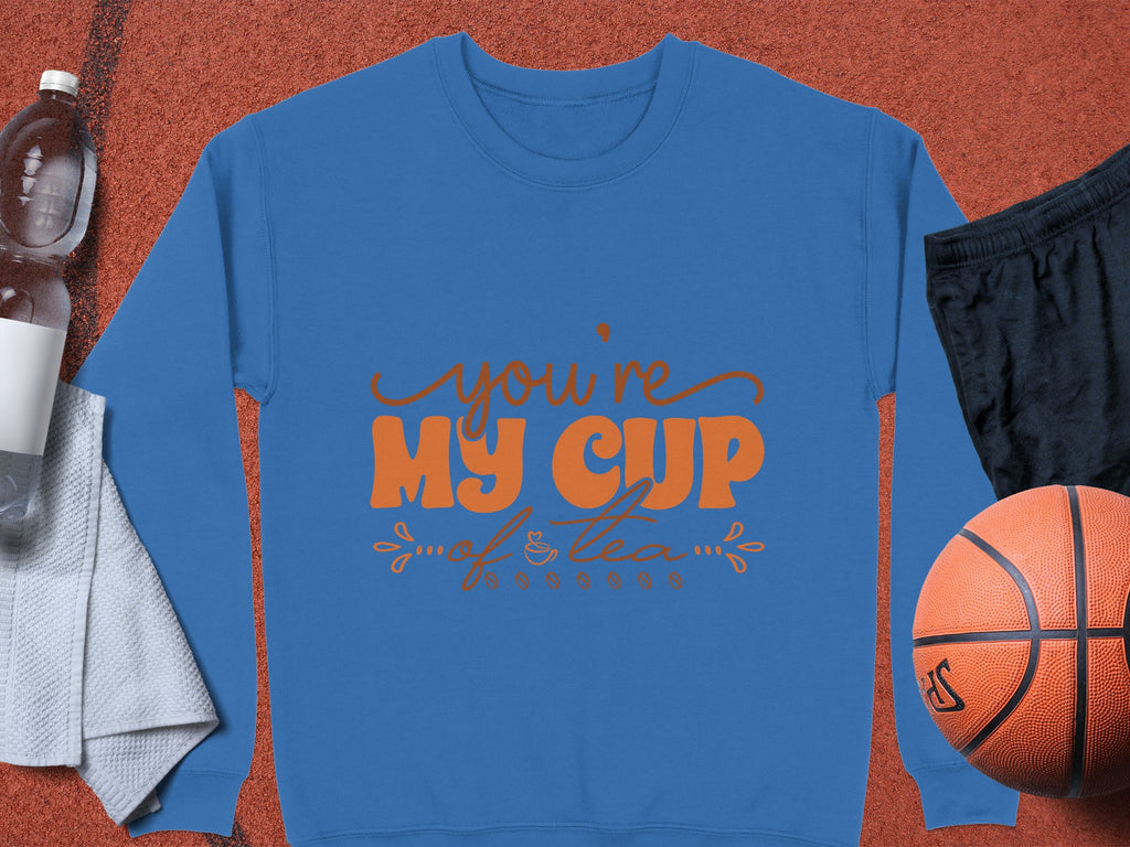 A cozy blue sweatshirt by Garment Graphics, with Youre My Cup of Tea in bright orange text, rests on a brown surface. Ideal for tea lovers, its accompanied by a basketball, black shorts, water bottle, and white towel.