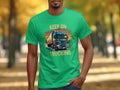 A person wearing a vibrant green Garment Graphics T-shirt with a detailed truck illustration and KEEP ON TRUCKING boldly above and below it, set against a blurred leafy backdrop—a perfect must-have for truck enthusiasts.
