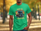 A person wearing a vibrant green Garment Graphics T-shirt with a detailed truck illustration and KEEP ON TRUCKING boldly above and below it, set against a blurred leafy backdrop—a perfect must-have for truck enthusiasts.