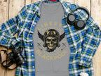A Garment Graphics gray T-shirt embodies the biker vibe with a skull with wings and goggles, featuring Liberty Motorcycles Blackpool Specialist 2023. It sits on a wooden surface beside a blue plaid shirt, camera, and headphones.