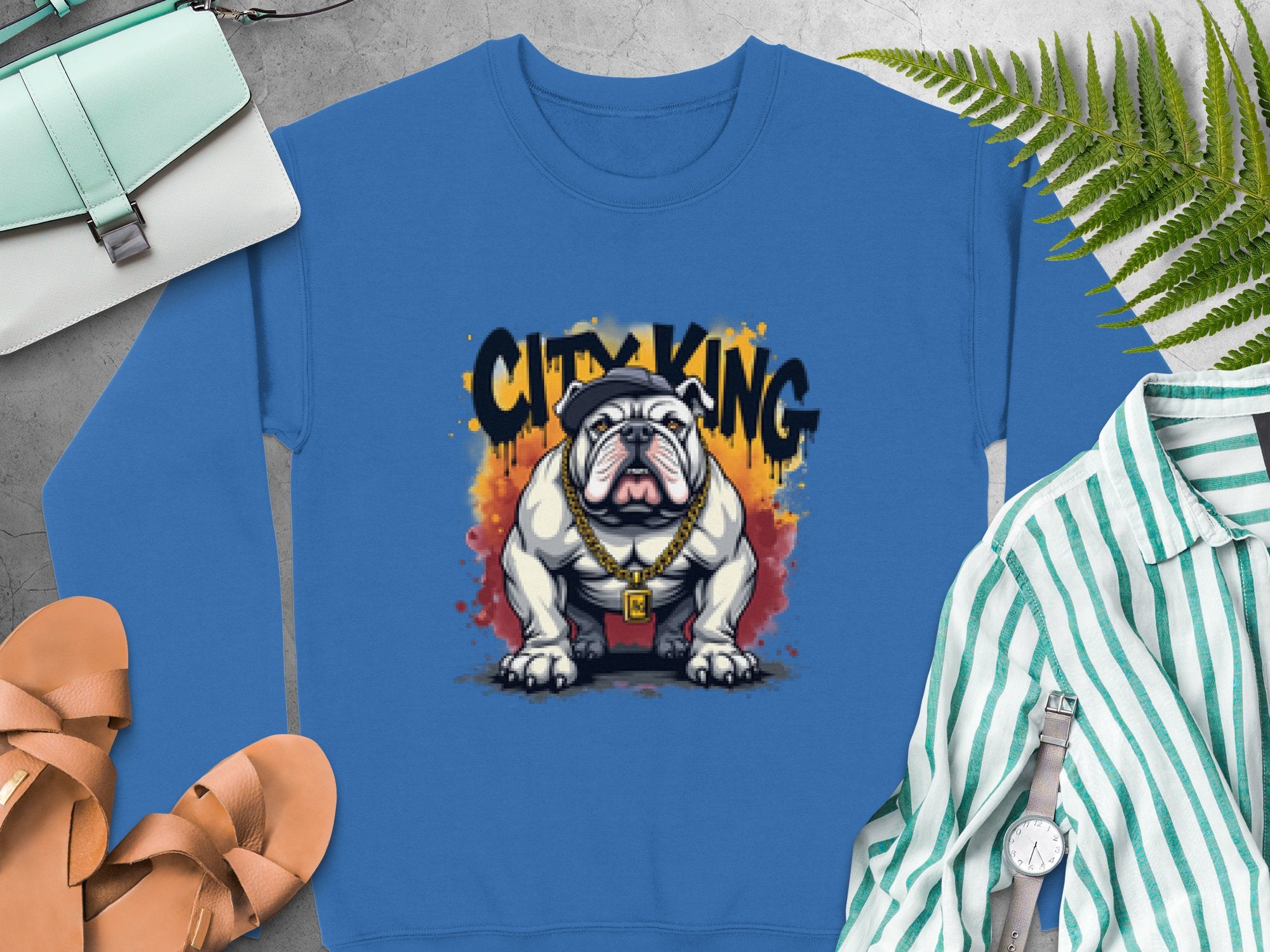 The Garment Graphics Bulldog Sweatshirt features a white bulldog graphic wearing a gold chain and red collar, topped with CITY KING in black graffiti-style text. Perfectly paired with sandals, a bag, and a striped shirt.