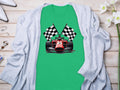 A Garment Graphics green T-shirt with a red racing car and crossed checkered flags is elegantly placed on light wood, paired with a gray cardigan, white high heels, scattered jewelry, and enhanced by white flowers for a charming motorsport-themed display.